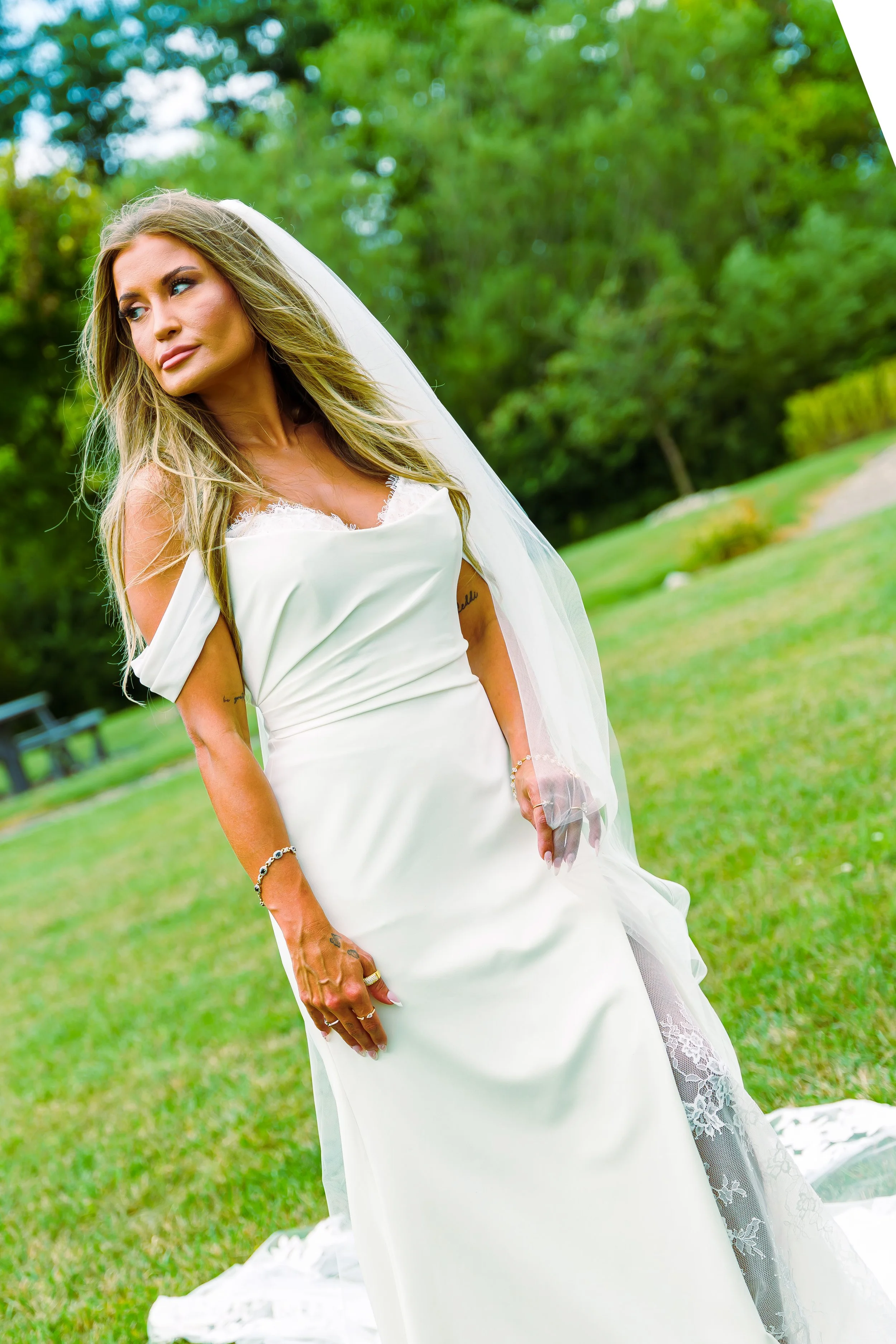 A woman in a white wedding dress with lace details, standing outdoors on a grassy area with green trees in the background, wearing jewelry and a veil.
