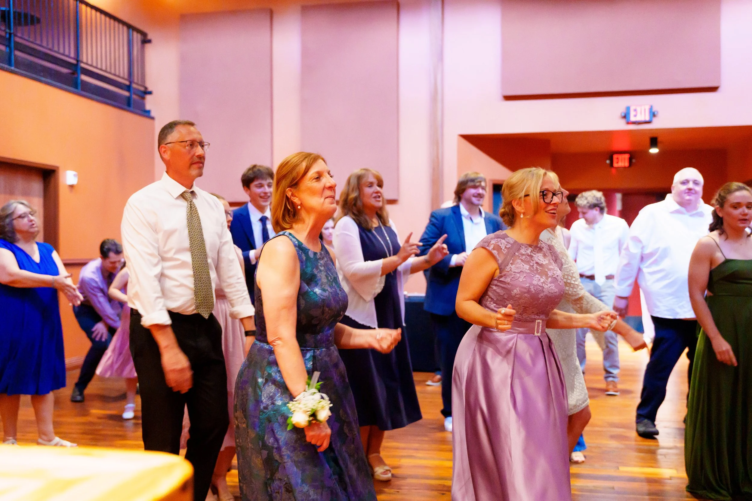 People dancing and celebrating at a wedding or formal event in a spacious indoor venue with pink and orange walls.