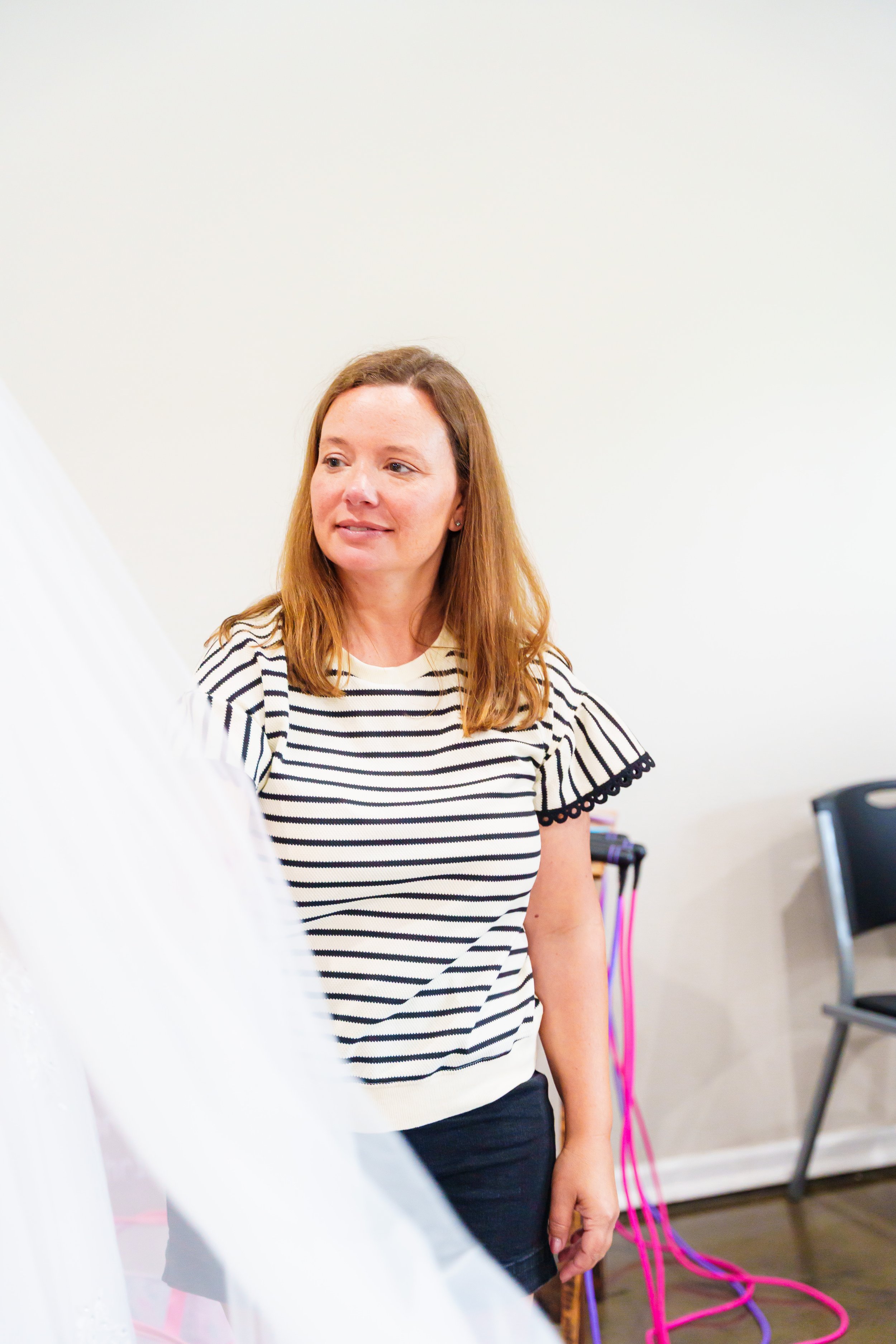 A woman with red hair wearing a black and white striped top, standing in a room with white walls. She is looking to her right with a slight smile. In the background, there are pink and purple cables out on the floor.