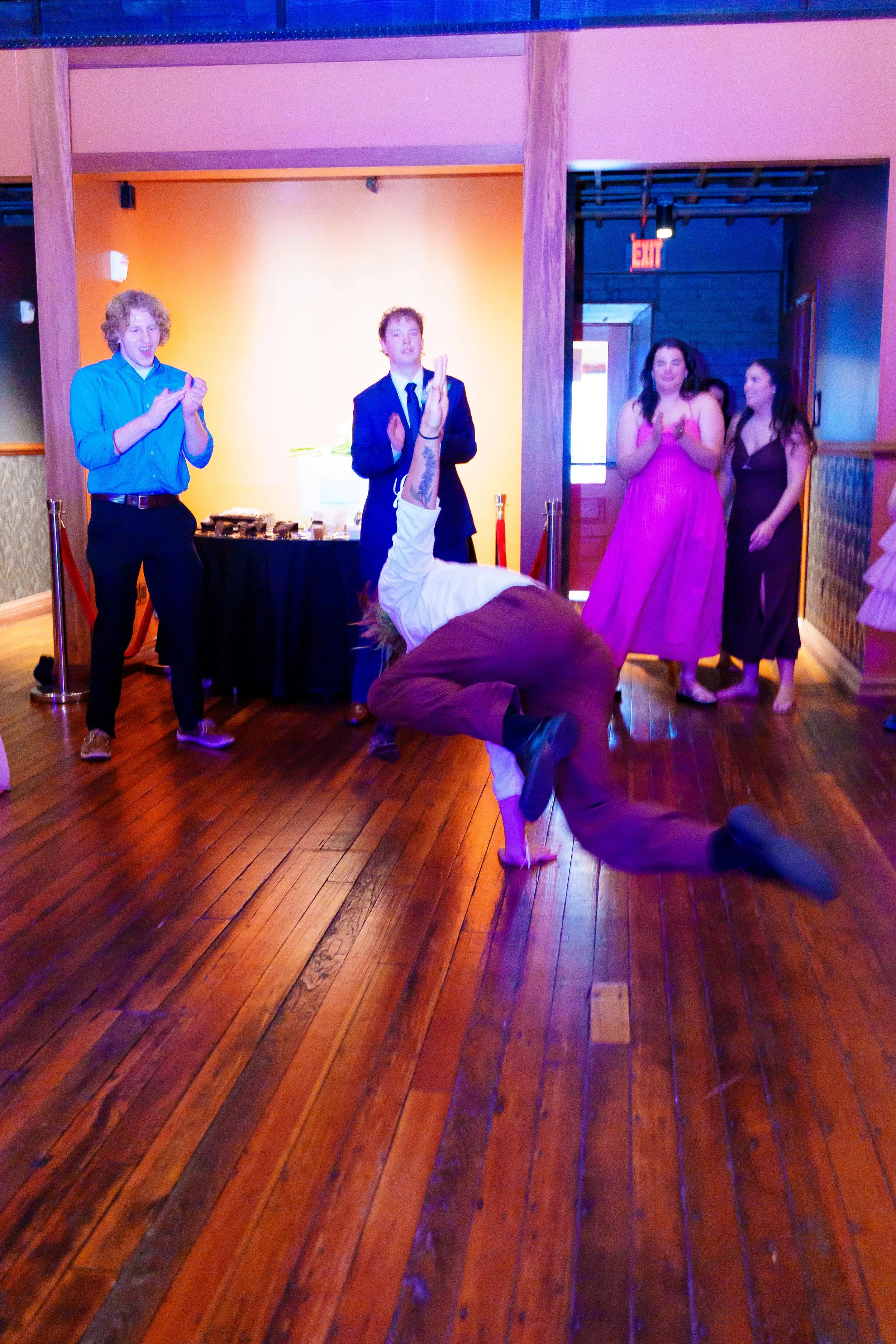 A group of people at a celebration or party, with some clapping and others watching a person in a brown jacket performing a breakdance move on the wooden floor, in a room with colorful lighting and people dressed in semi-formal attire.