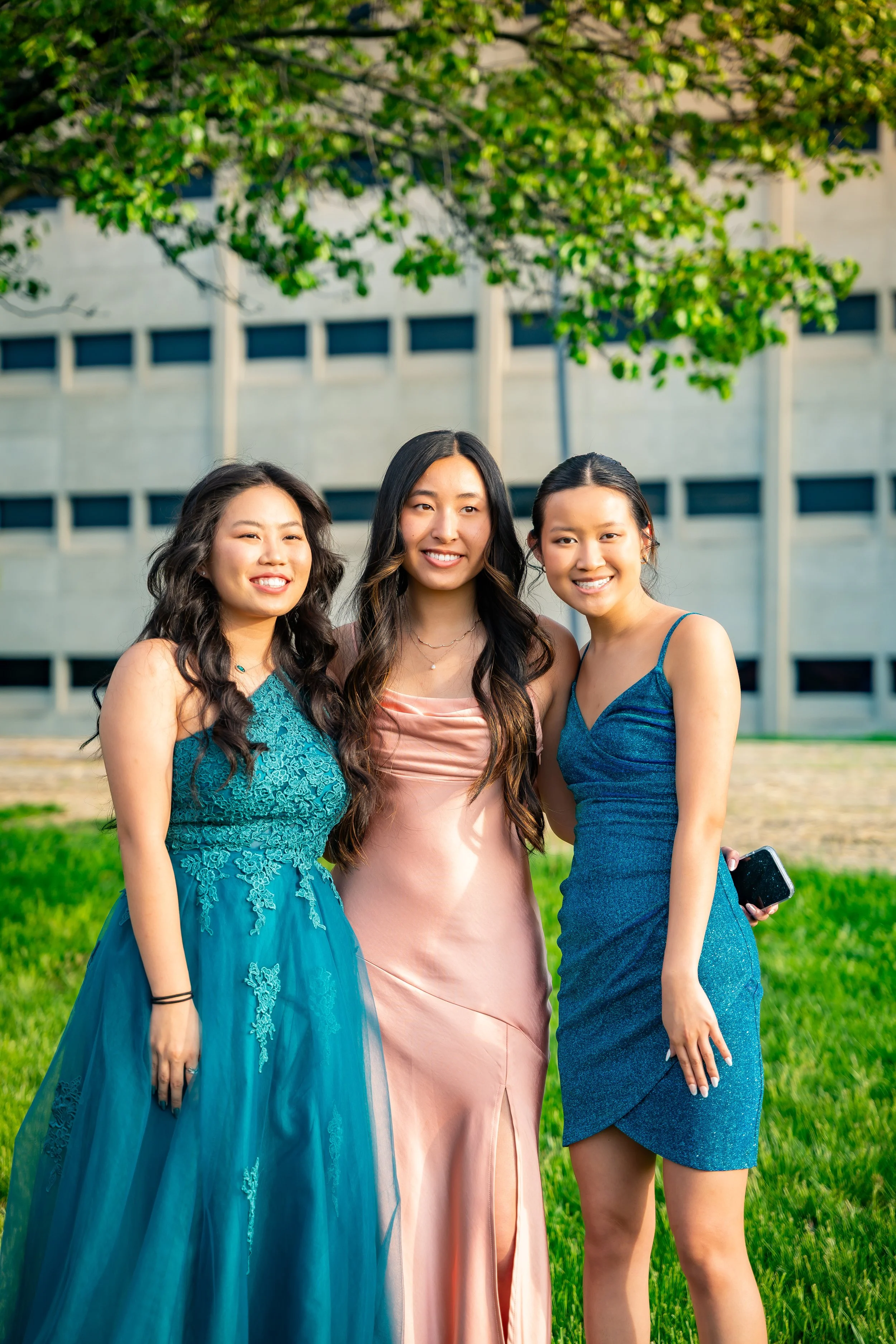 Three young women in elegant dresses standing outdoors on green grass with a building and trees in the background, smiling at the camera.