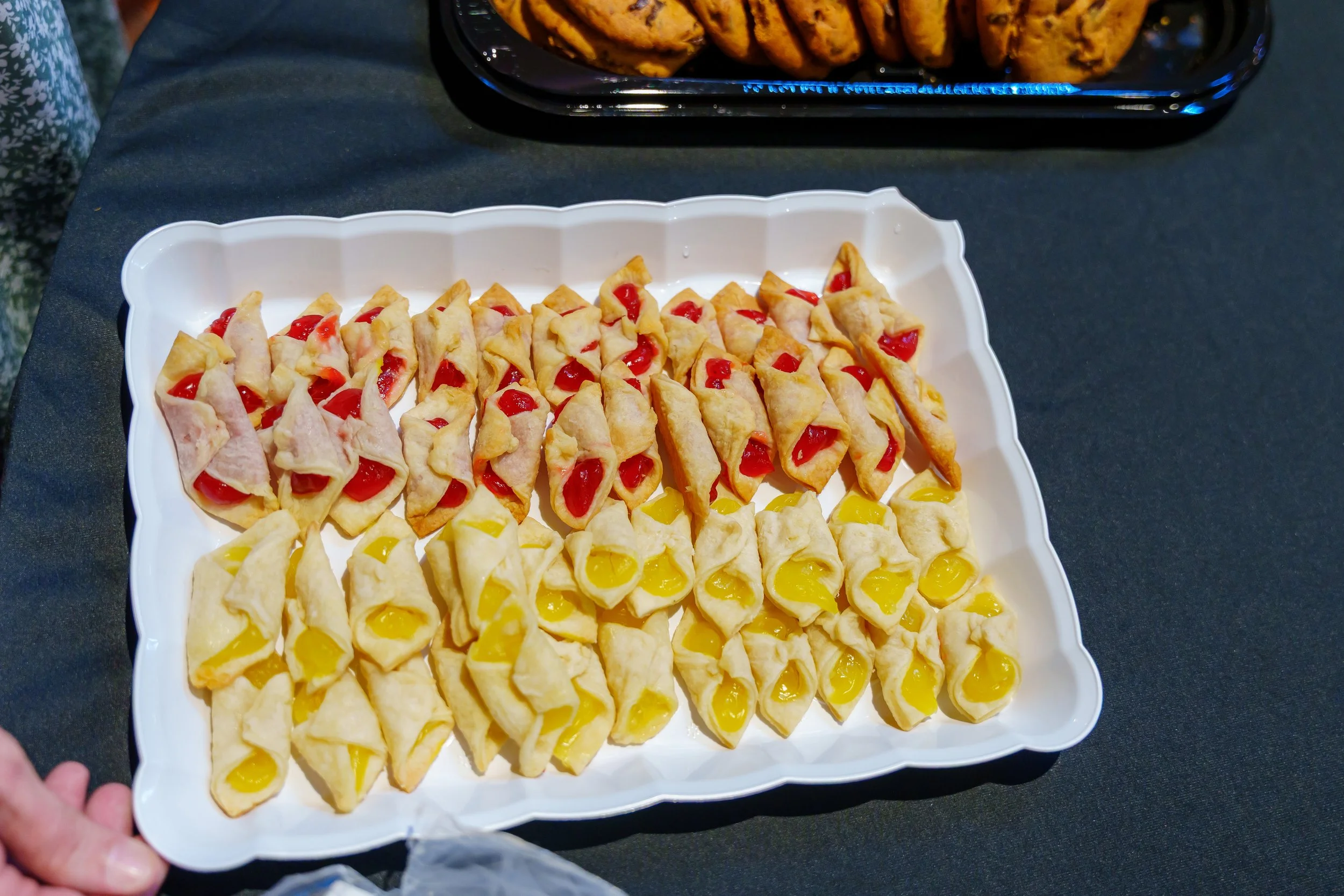 A tray of bite-sized pastries filled with yellow and red jelly, arranged in neat rows on a white platter, with a container of cookies visible in the background.