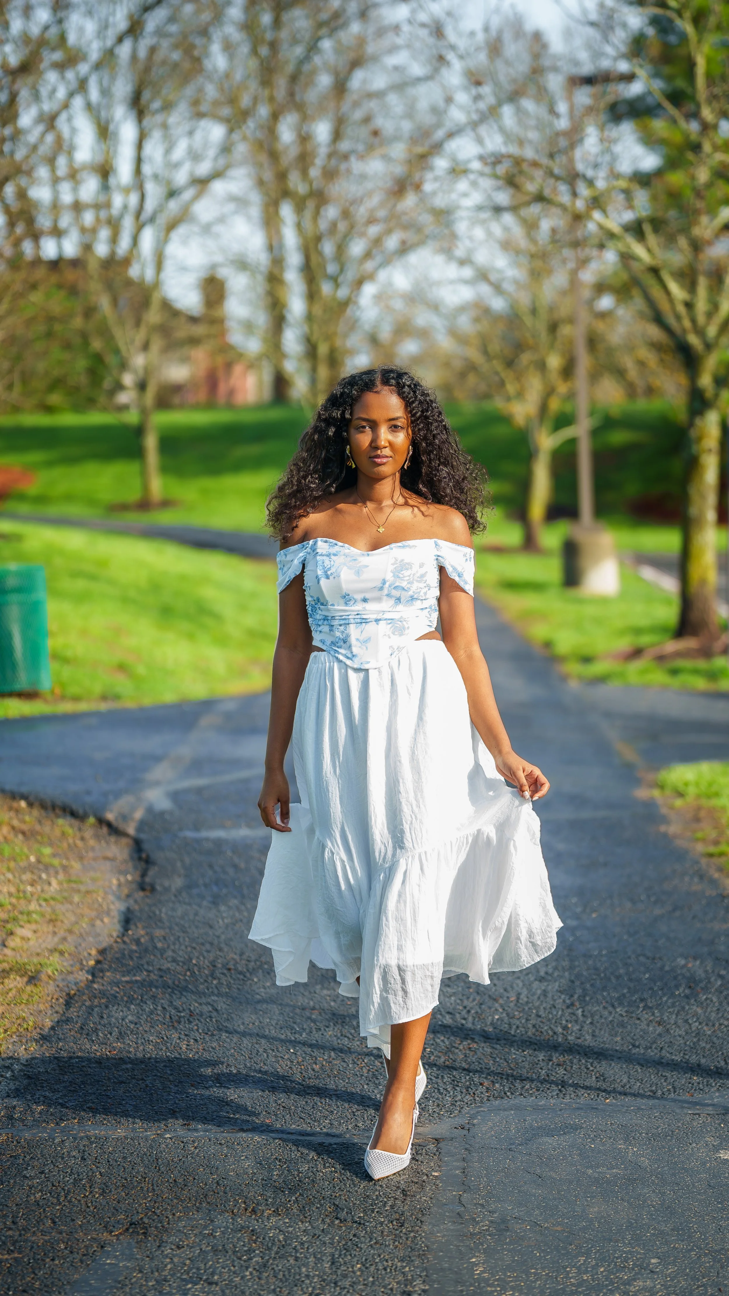 A woman walking on a paved path in a park, wearing a white dress and heels, with trees and greenery in the background.
