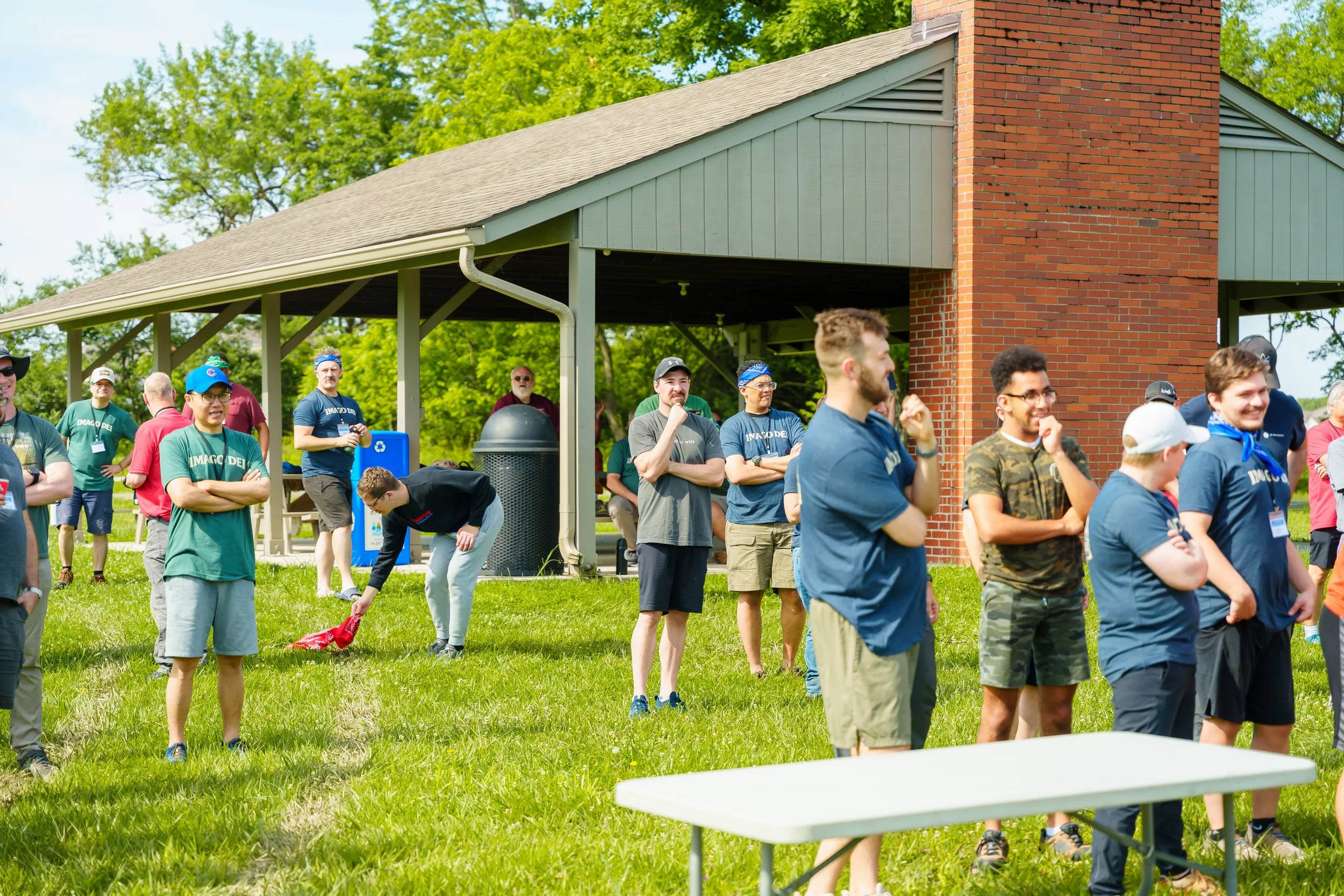 Group of people gathered outdoors on a grassy area near a pavilion with a brick chimney, some people are smiling and talking, others are standing with arms crossed or looking around.