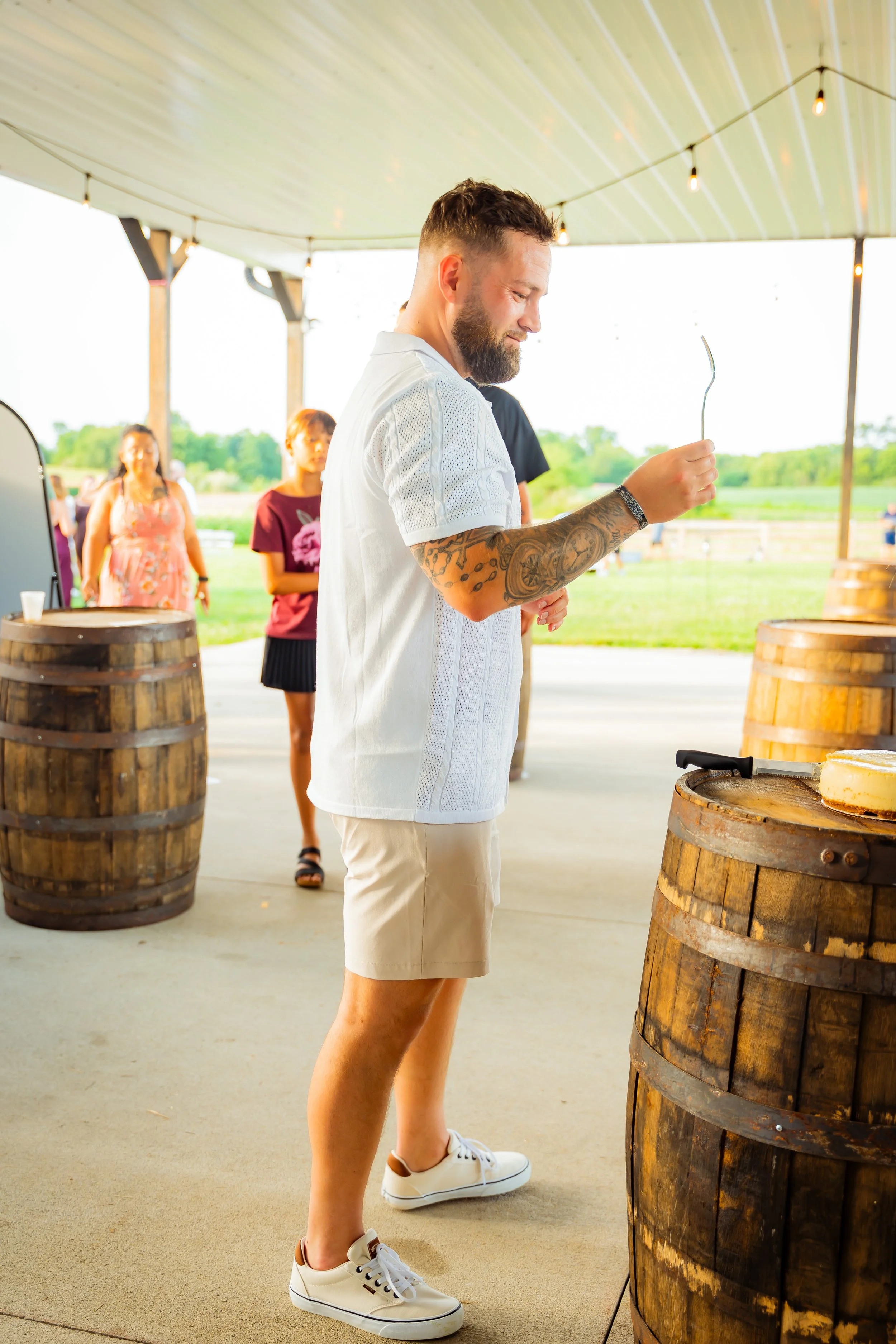 A man with a beard and tattoos on his arms, dressed in a white shirt, beige shorts, and white sneakers, is holding a cheese knife and appears to be cutting cheese on a barrel at an outdoor gathering. Two women are seen in the background, one in a flo