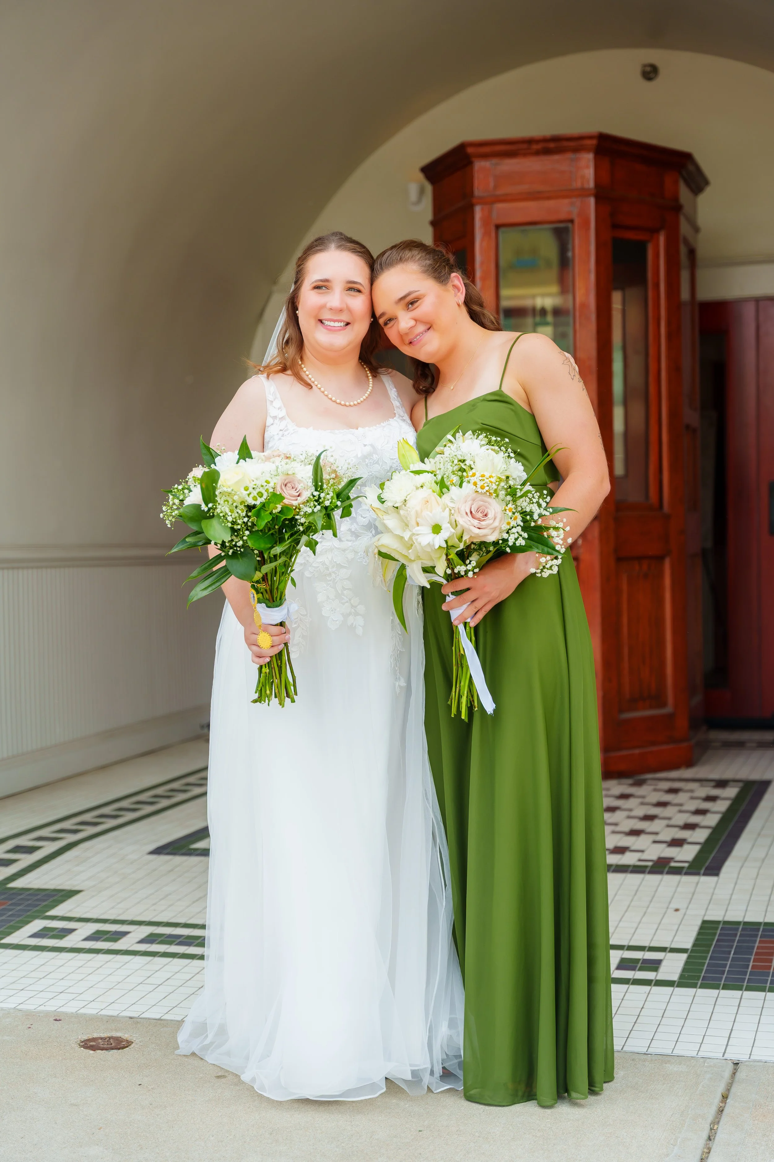 Two women in wedding attire holding bouquets, standing close together and smiling inside a building with a tiled floor and wooden cabinet in the background.