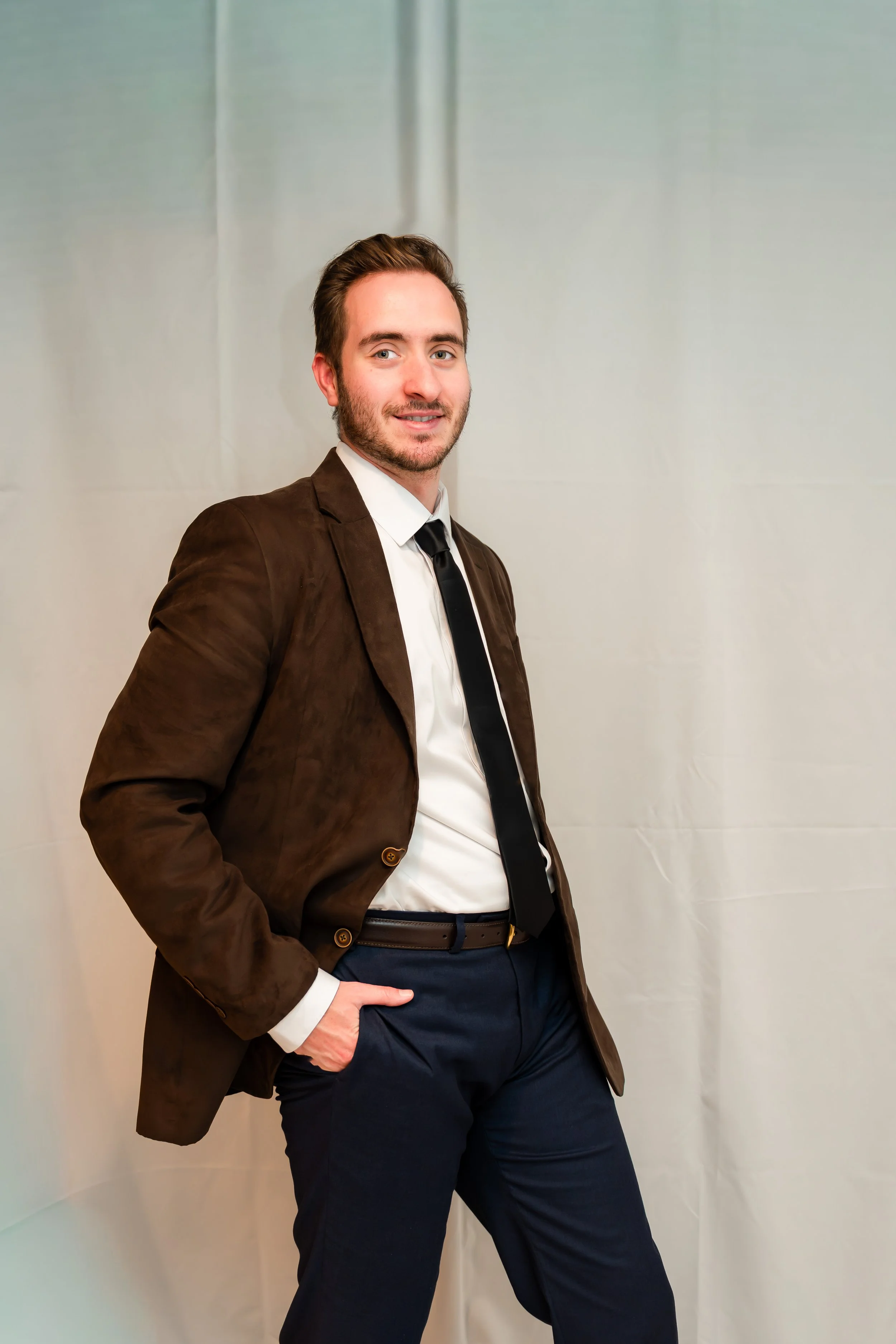 A man dressed in a brown blazer, white shirt, and black tie, with navy pants, posing confidently with his right hand in his pocket and leaning slightly against a plain light-colored wall.