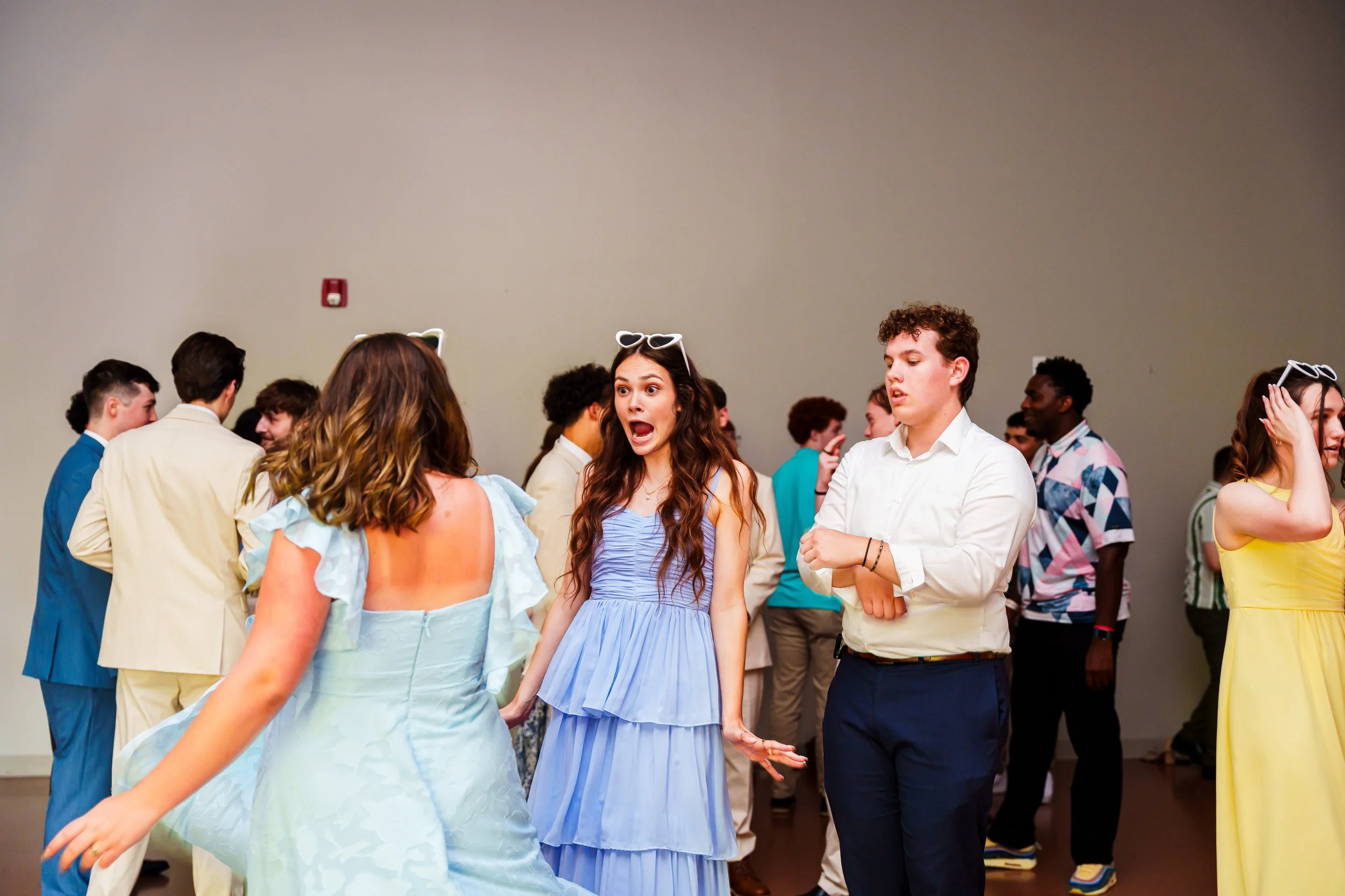A group of young people dancing at a social event, dressed in colorful and semi-formal attire, with a plain beige wall background.