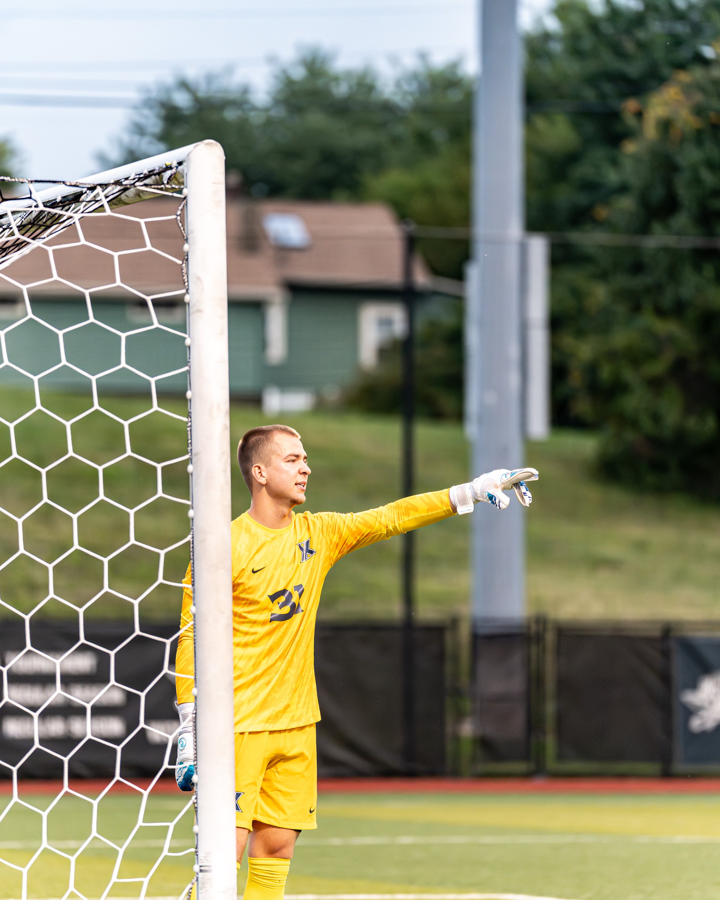 A male soccer goalkeeper in a yellow uniform standing near the goalpost, extending his arm and pointing during a game on a grassy field.