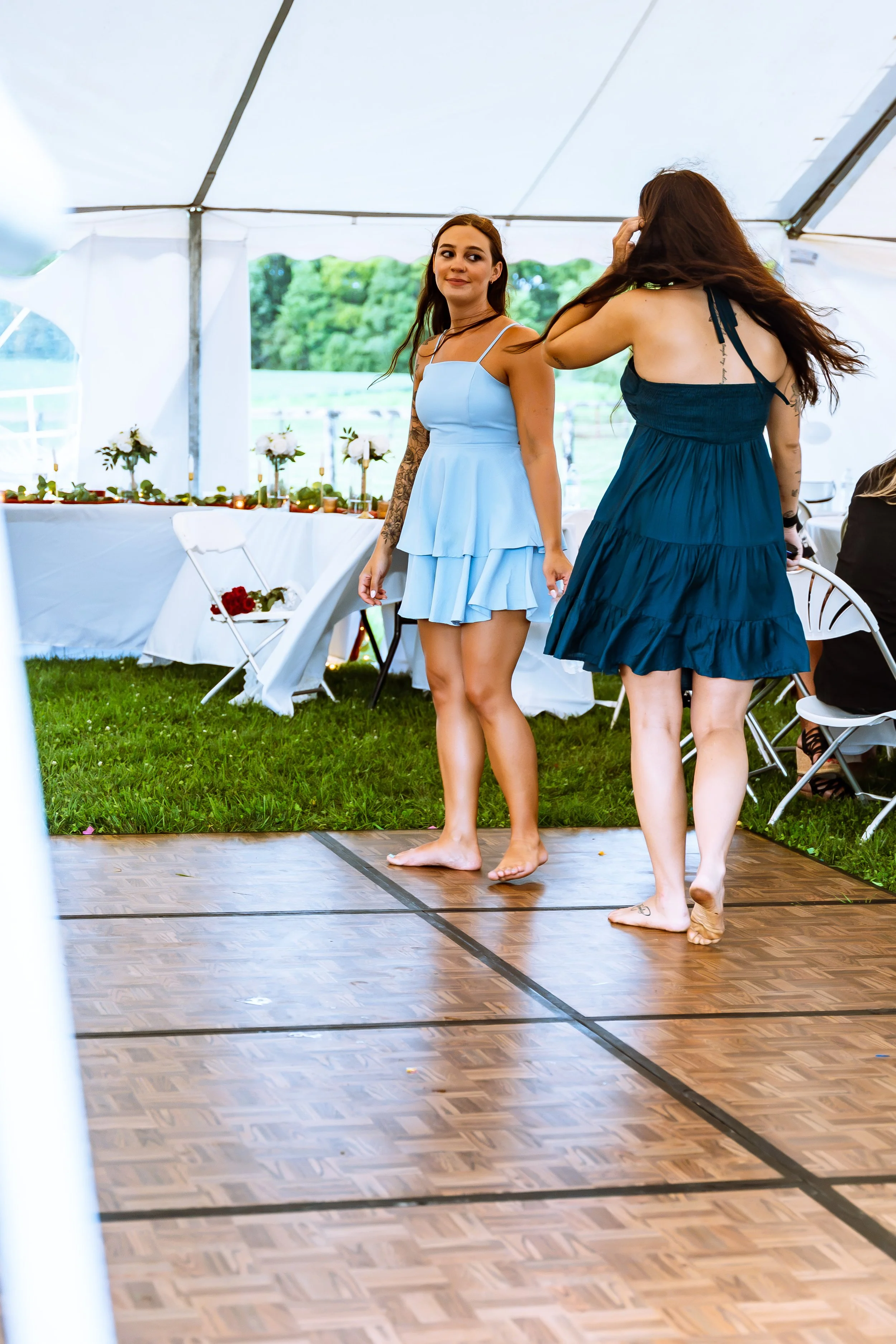 Two women are barefoot and standing on a small dance floor inside a white tent. One woman in a light blue dress is looking back and smiling, while the other woman in a dark blue dress has her back to the camera and is touching her hair. In the backgr