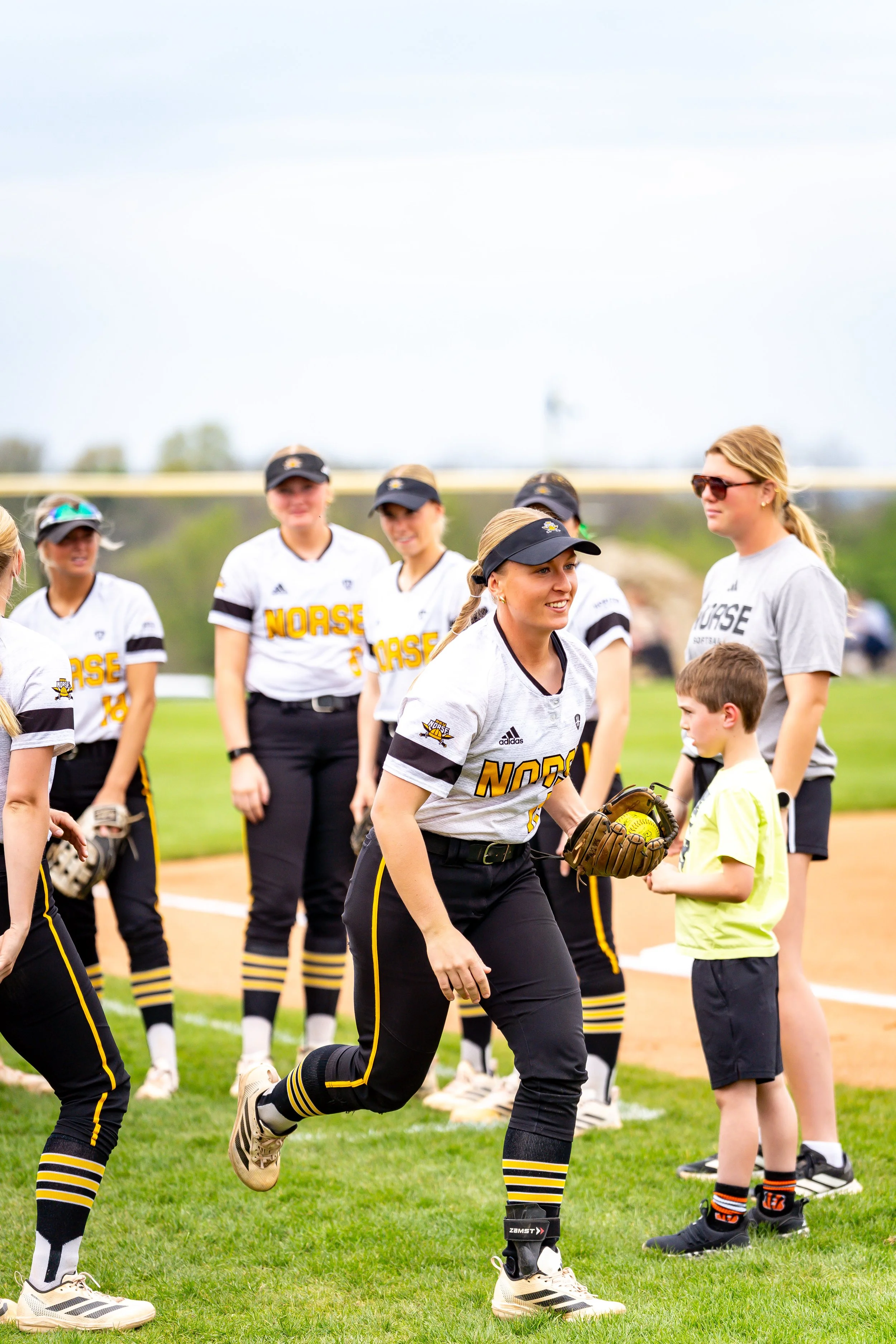 Group of female softball players in uniform on a field, some wearing caps and sunglasses, with a woman in the foreground running with a glove, and children standing nearby.