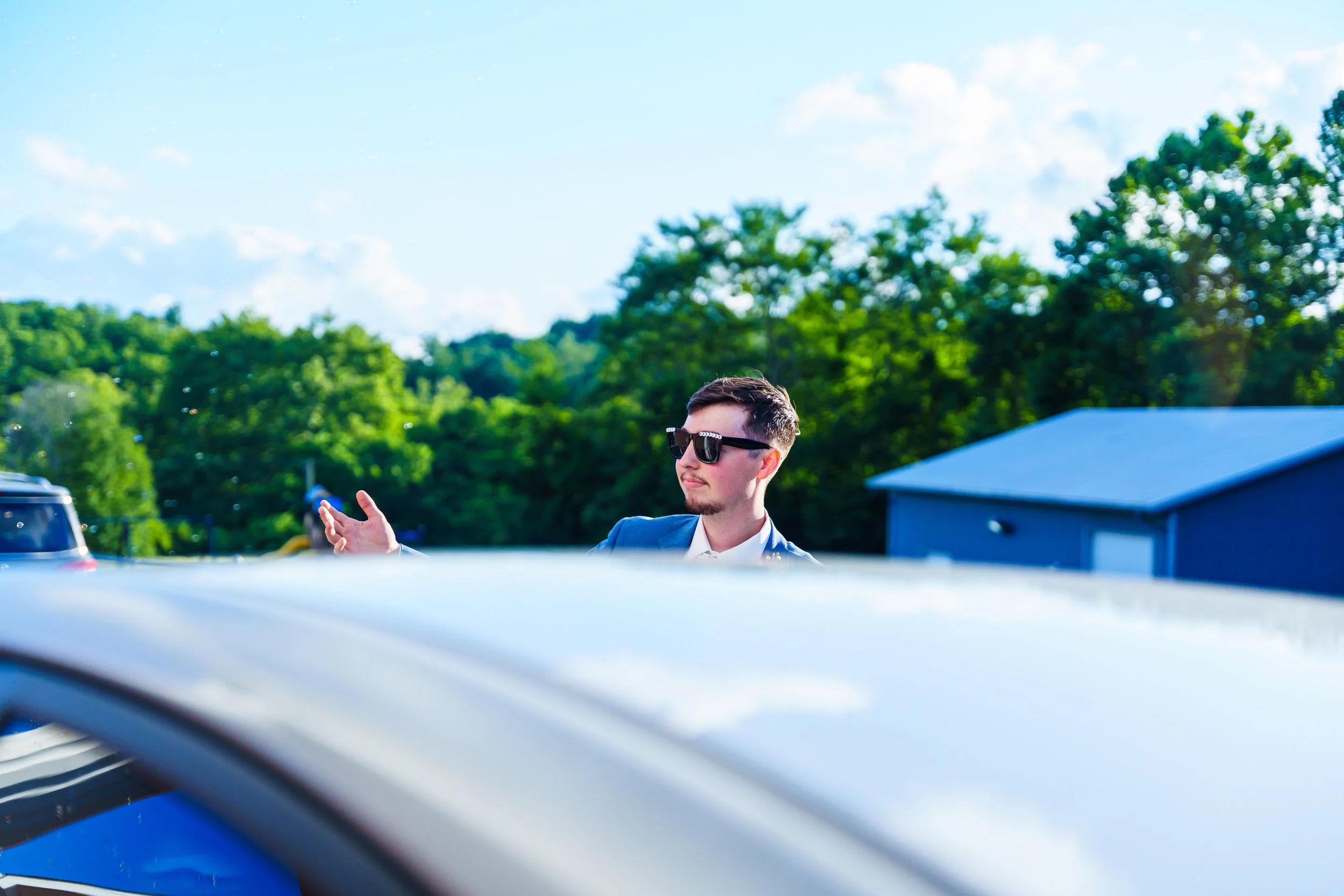 A young man with short dark hair, wearing sunglasses and a dark blazer, is standing outdoors in front of a green, leafy landscape under a partly cloudy blue sky.
