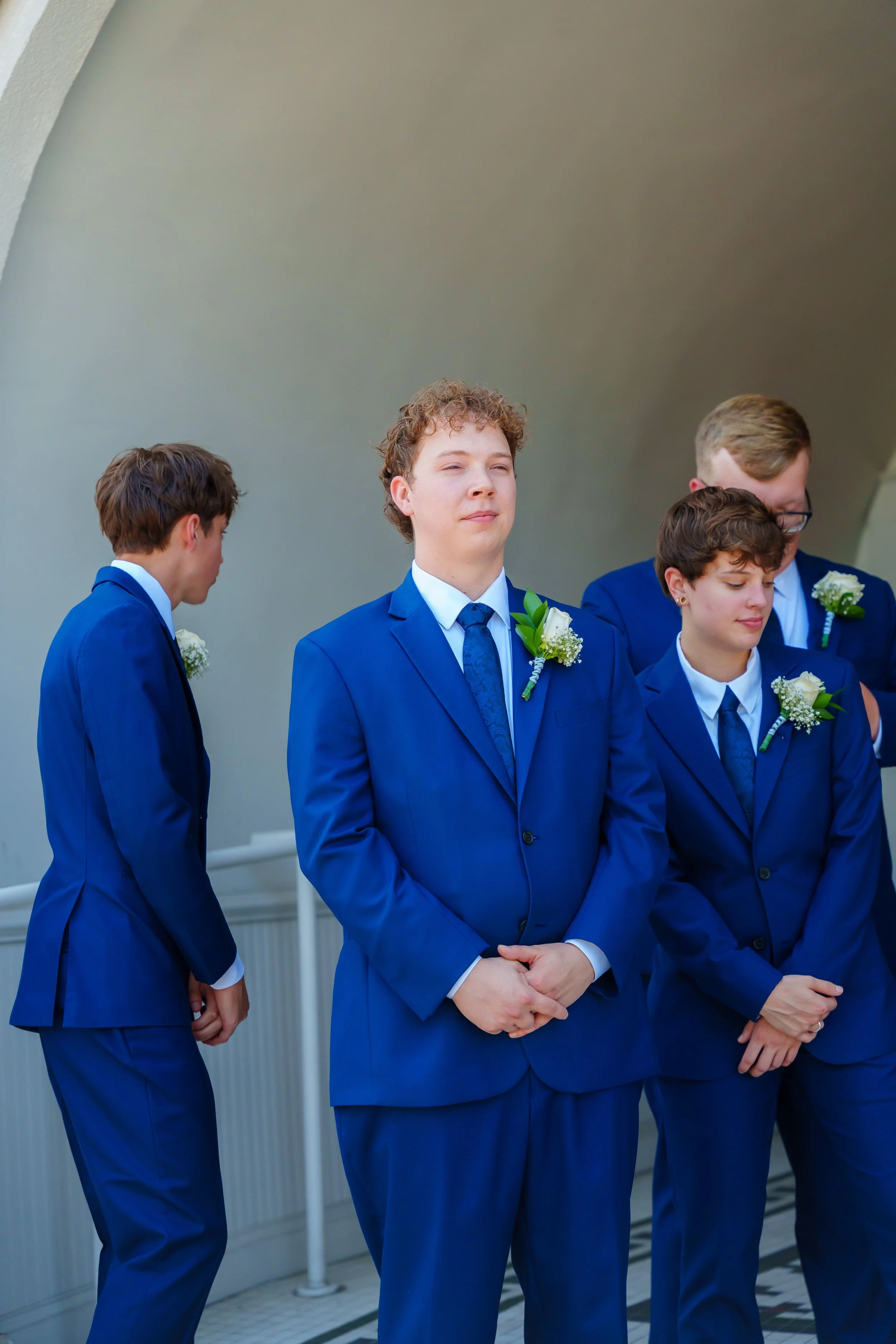 Four young men in blue suits wearing white shirts, dark blue ties, and white boutonnieres standing outdoors near a building with a white railing.