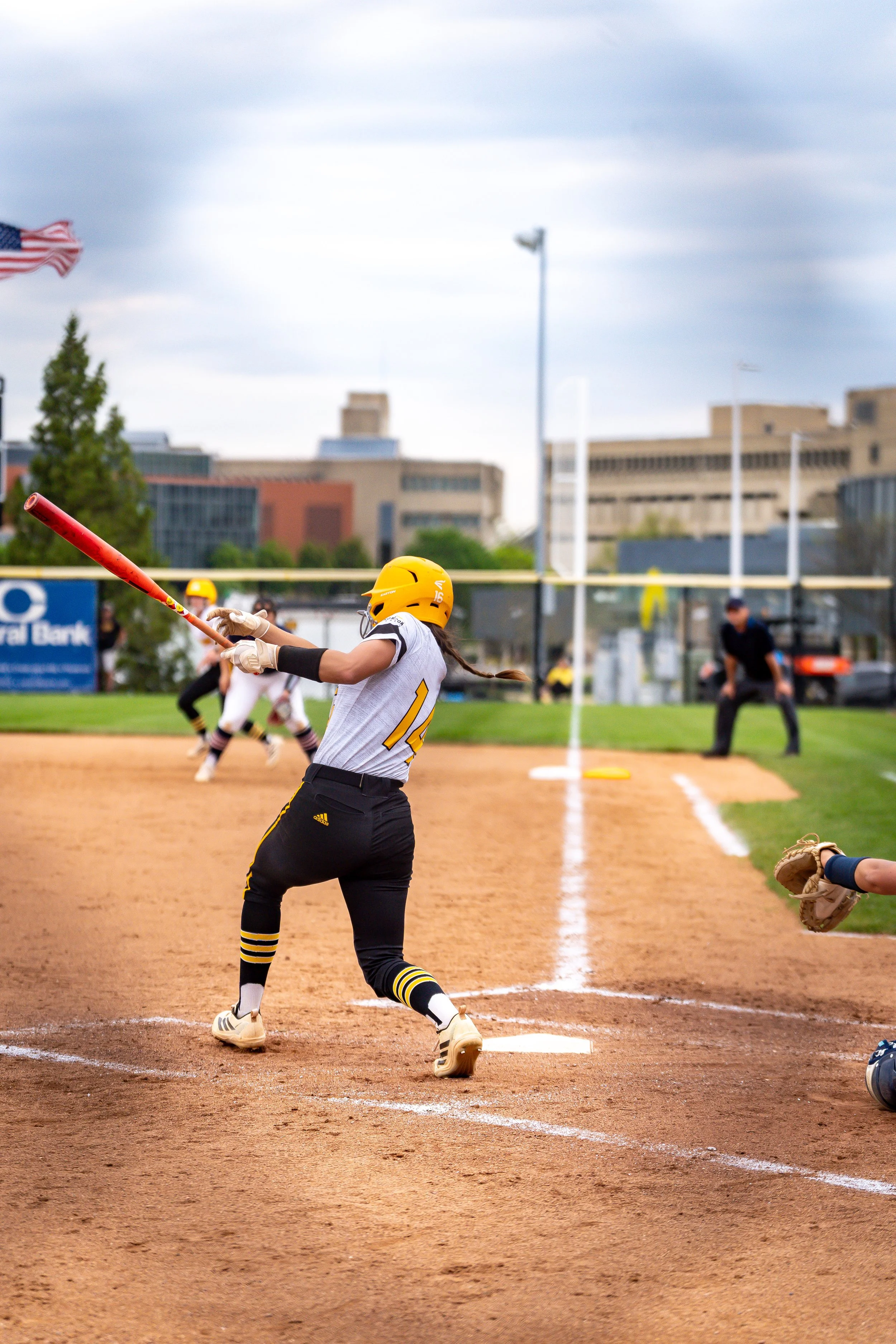 A female softball player in a yellow helmet and black pants is running towards first base on a softball field during a game, with other players and an umpire visible in the background.