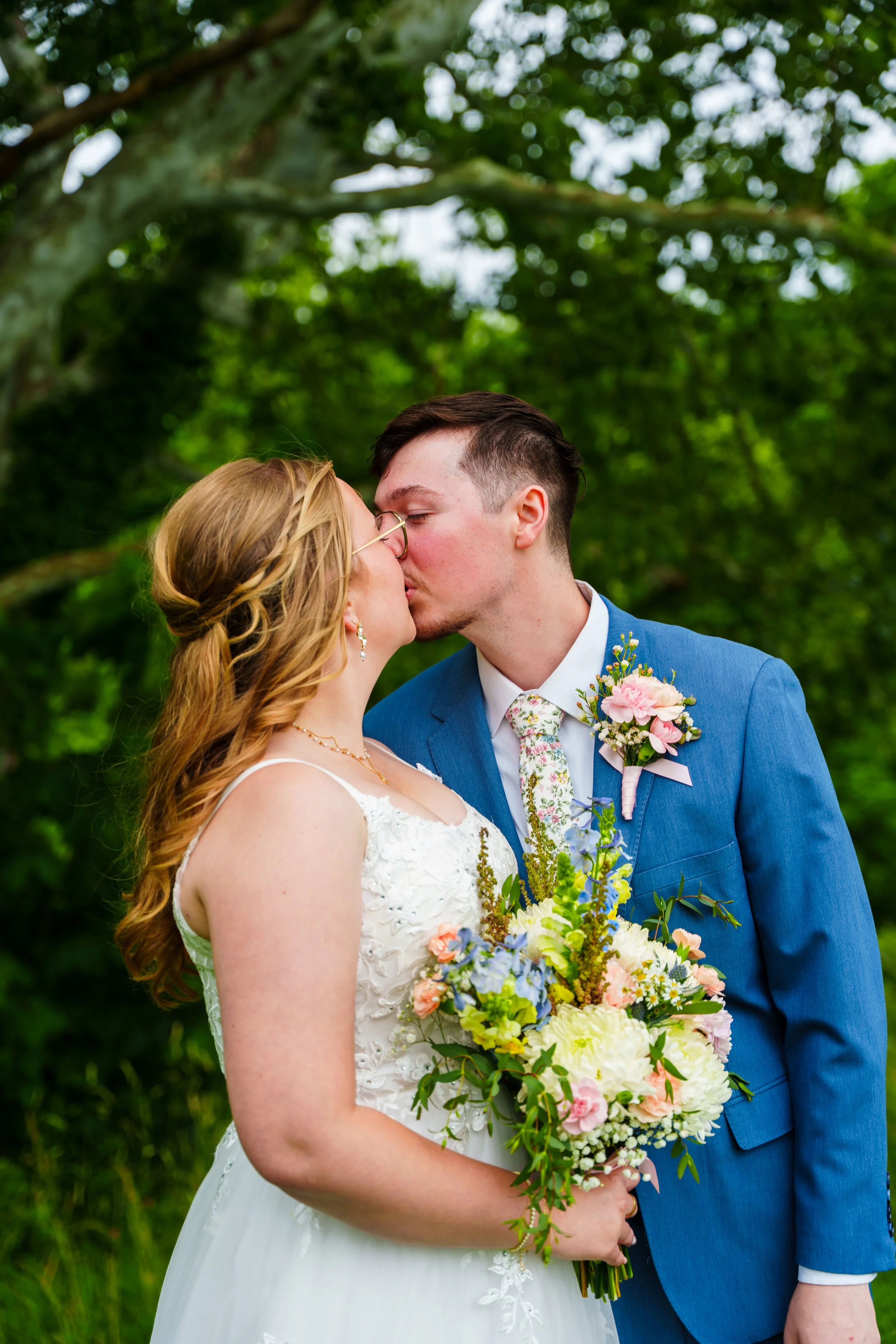 A bride and groom kiss outdoors on their wedding day. The bride has red hair, wears glasses, and a white dress with floral embroidery. The groom has dark hair, a beard, and is dressed in a blue suit, white shirt, and floral tie, with a boutonniere. T