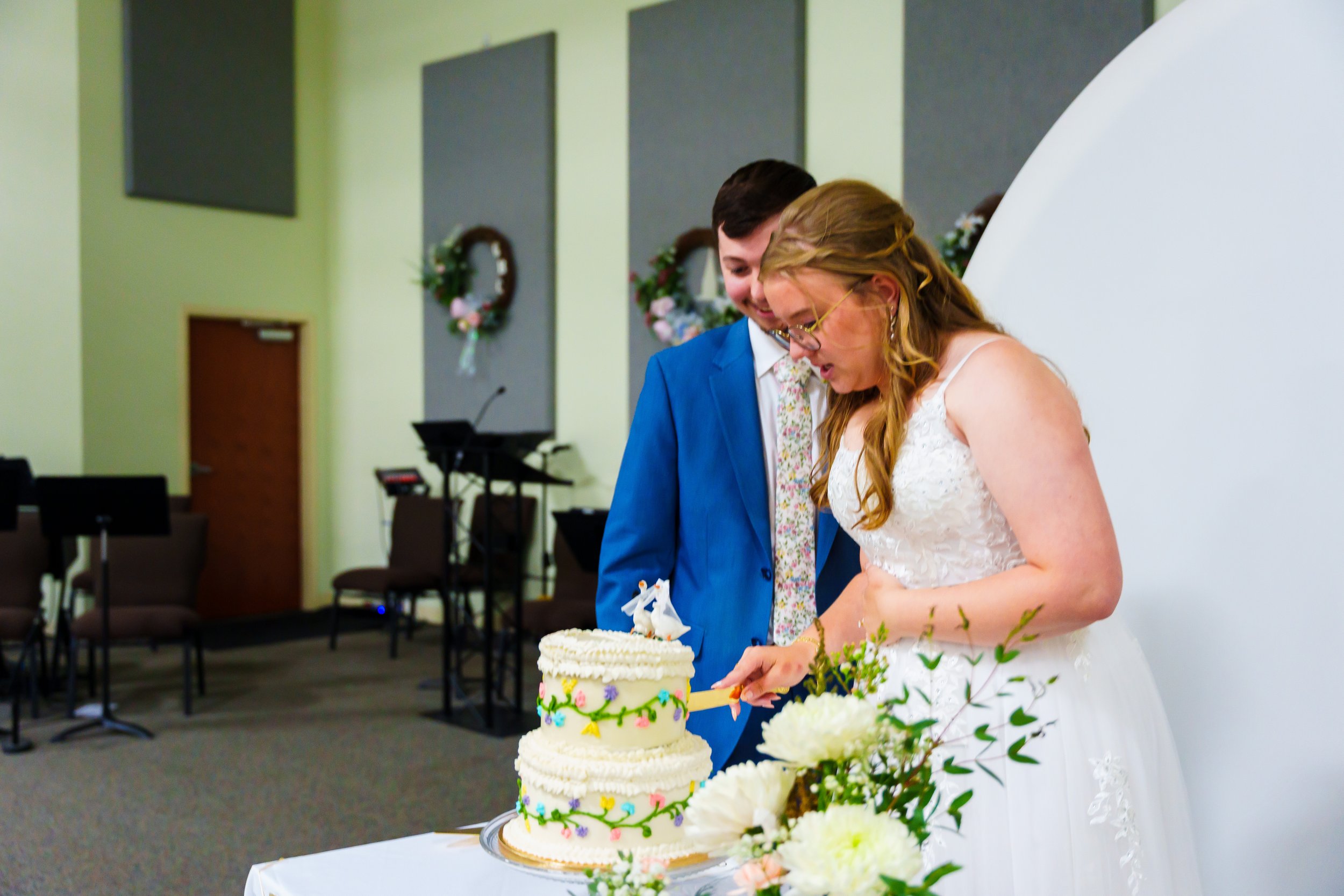 A bride and groom cutting a wedding cake at a reception, decorated with colorful flowers and greenery, with floral wreaths hanging on the wall behind them.