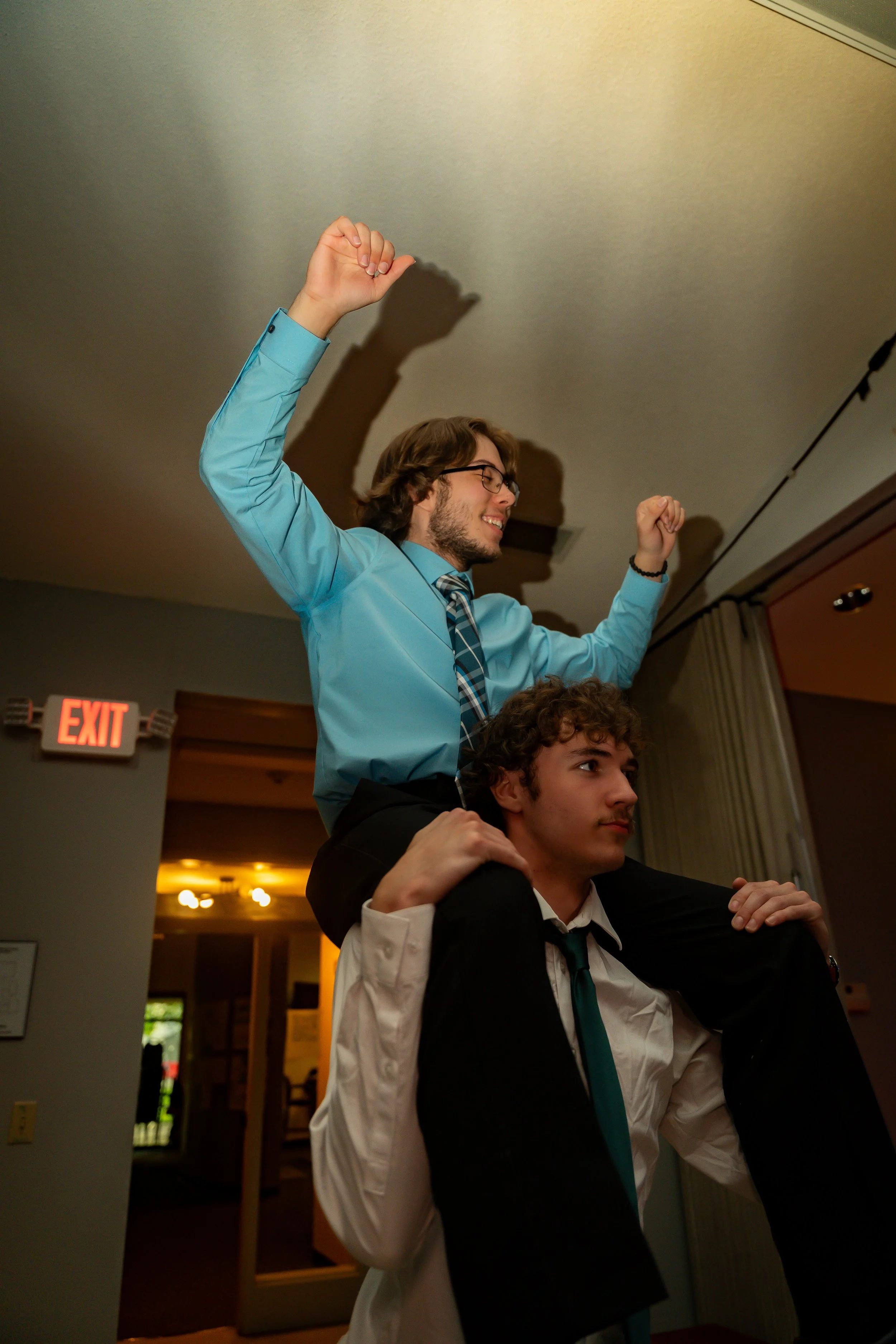 Two young men, one with glasses and a blue shirt, standing behind the other who is wearing a white shirt and a black vest, in an indoor setting with an exit sign visible in the background. The man in the blue shirt is smiling and raising both arms in