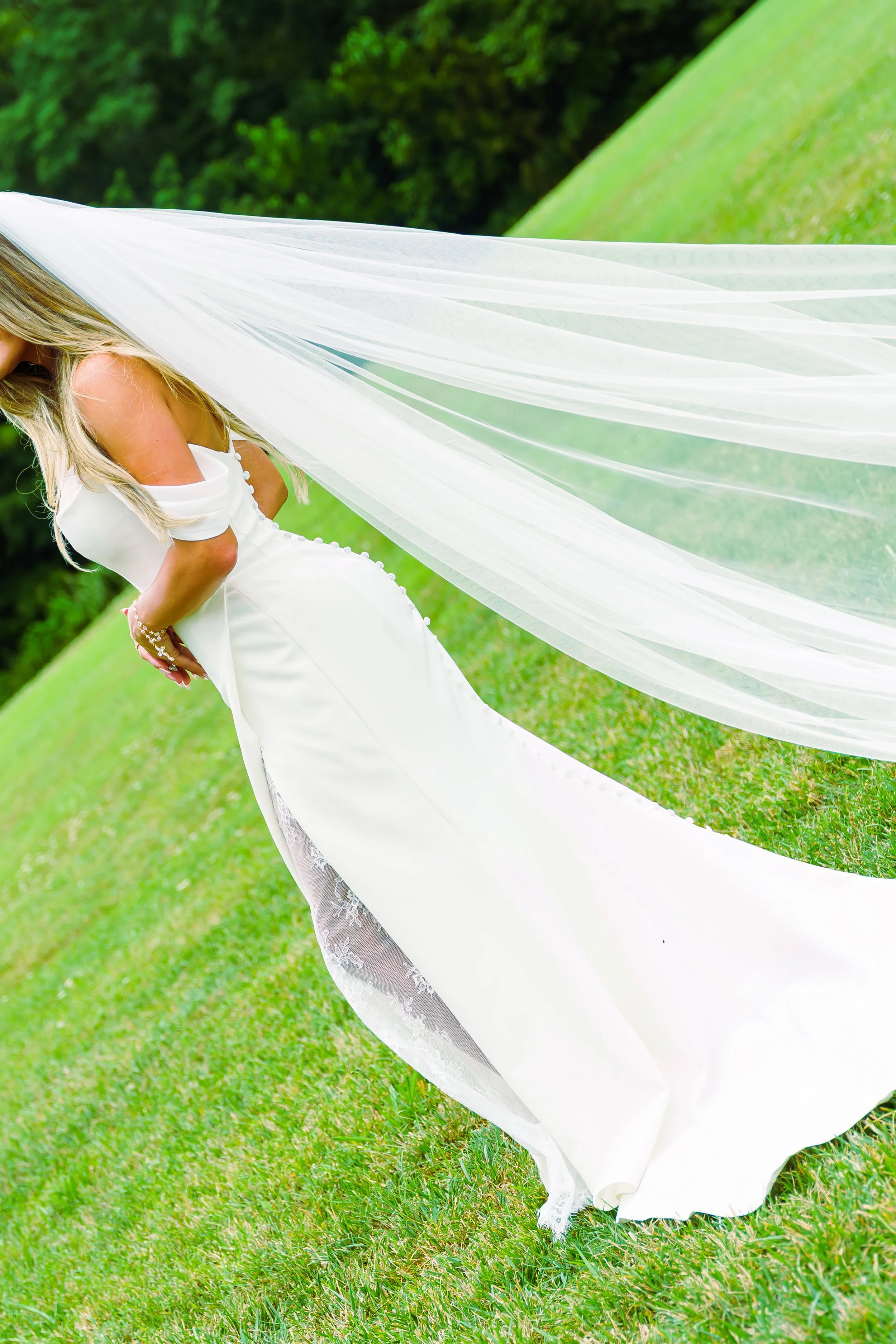 A woman in a white dress with lace details standing on green grass with lush trees in the background, holding a long white sheer fabric overhead.