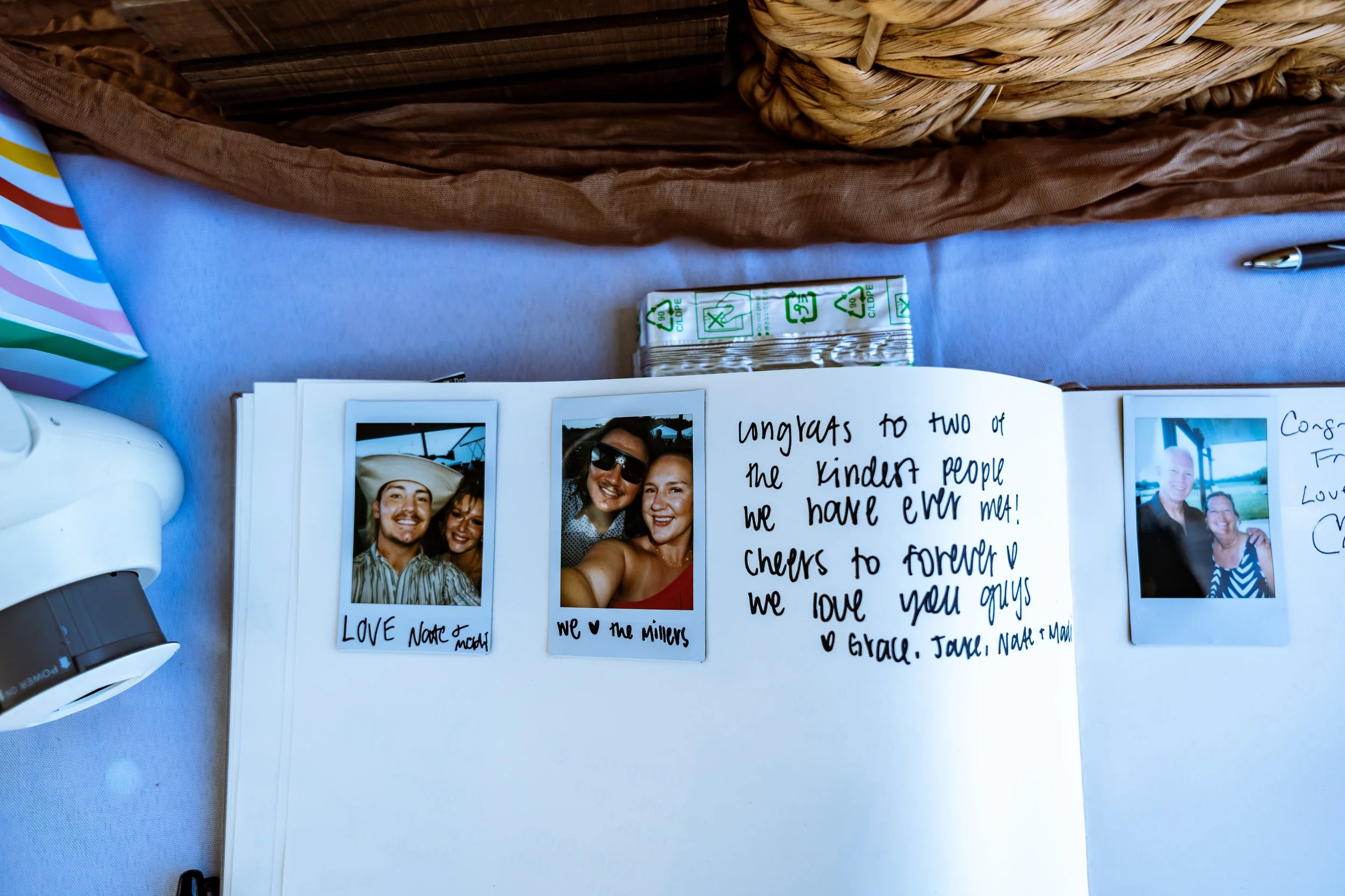 Open scrapbook with Polaroid photos and handwritten notes, placed on a table with a pen, notepad, and a basket in the background.