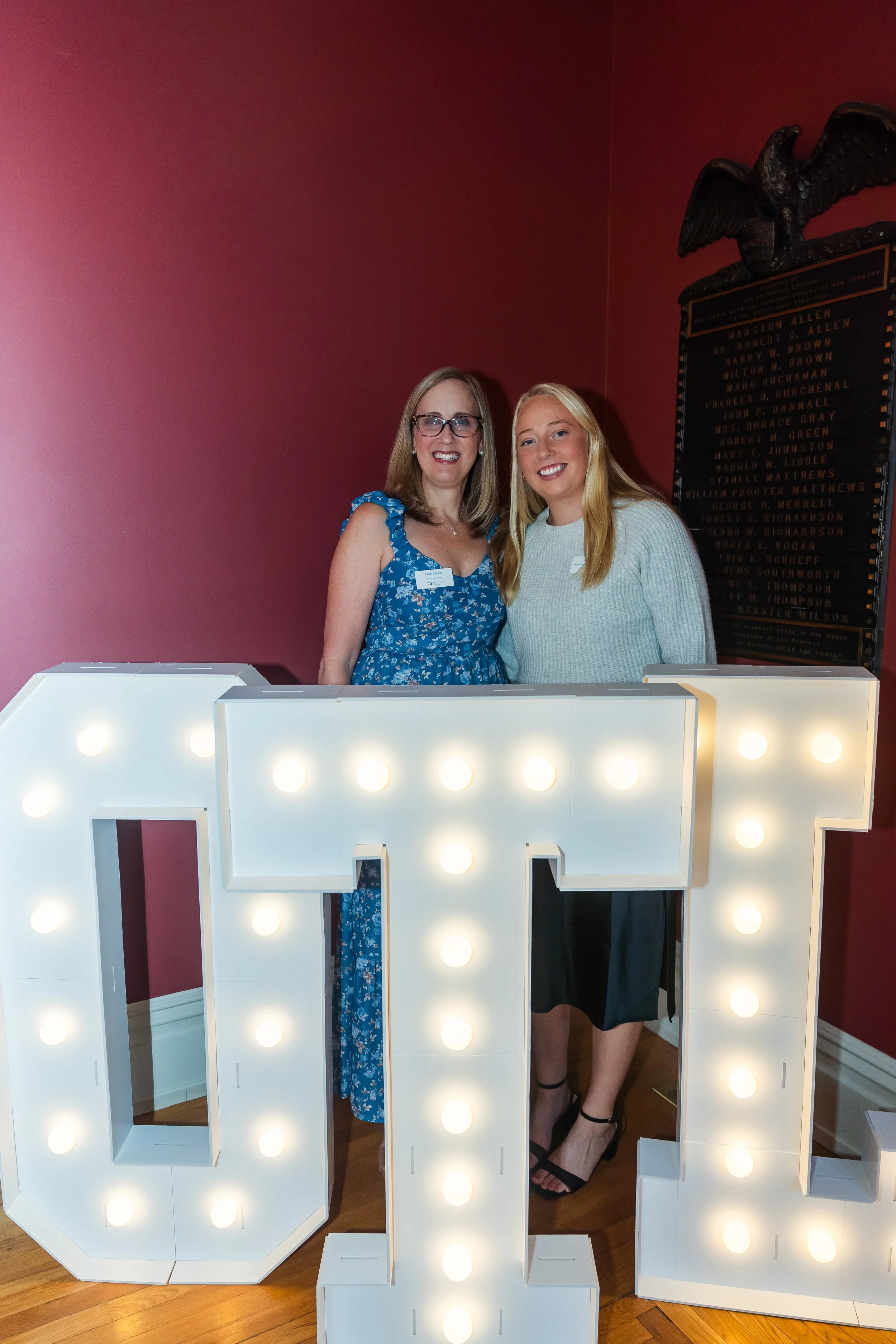 Two women standing behind a large white marquee letter 'H' with round lights, in a room with red walls and a wood floor. One woman wears a blue floral dress and glasses; the other wears a gray sweater and black skirt.