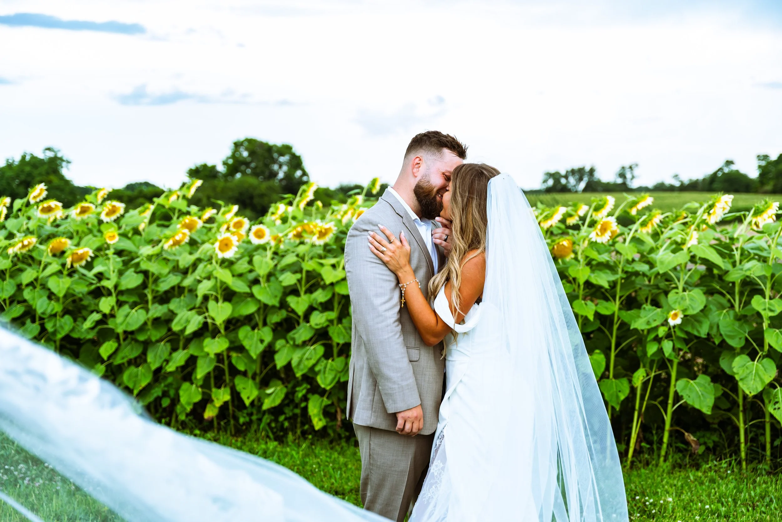 Newlywed couple sharing a kiss in a sunflower field on a cloudy day.
