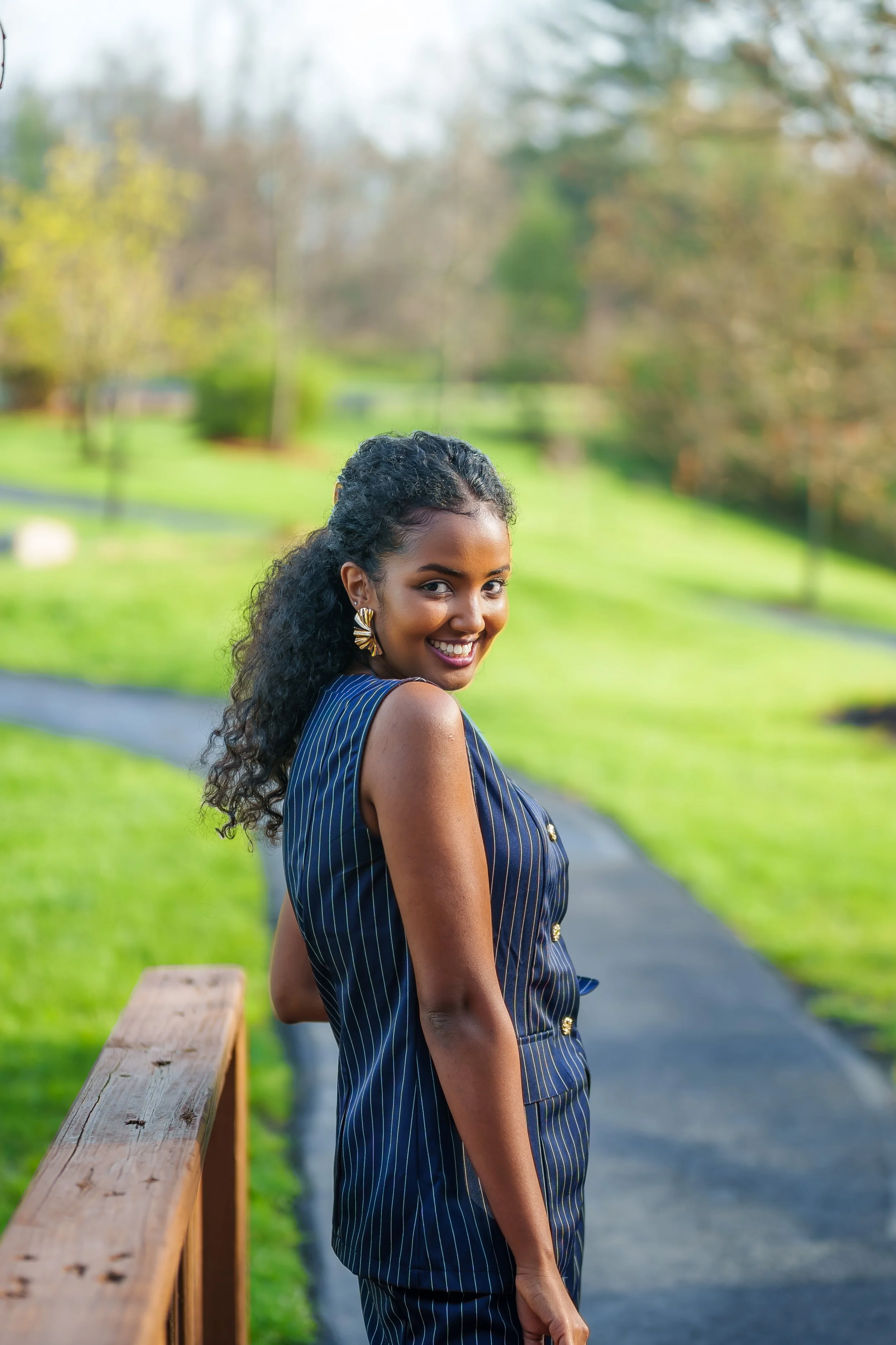 A smiling woman with curly black hair, wearing a navy blue pinstripe sleeveless blazer, standing outdoors on a park trail with green grass and trees in the background.