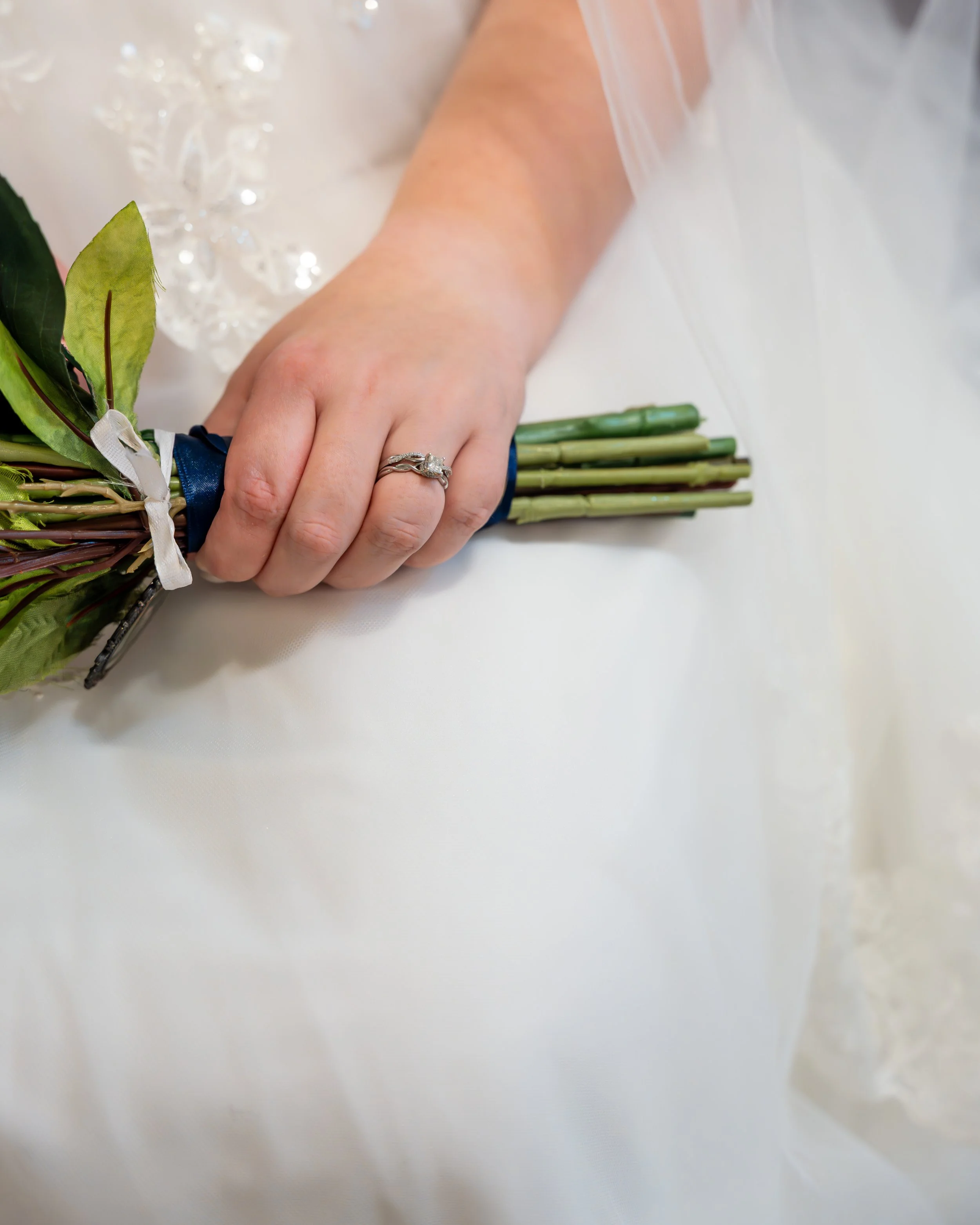 Close-up of a bride's hand holding a small bouquet of green leaves and stems with a blue ribbon, showing a wedding ring on her finger.