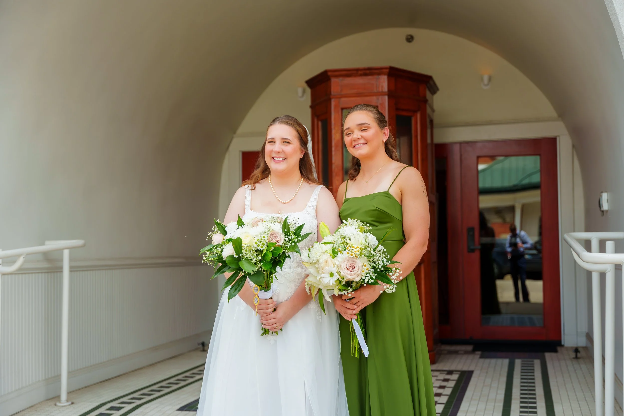 Two women, one in a white wedding dress holding a bouquet of white and pink flowers, and the other in a green dress also holding a bouquet, standing inside a building with an arched ceiling and a wooden cabinet in the background.