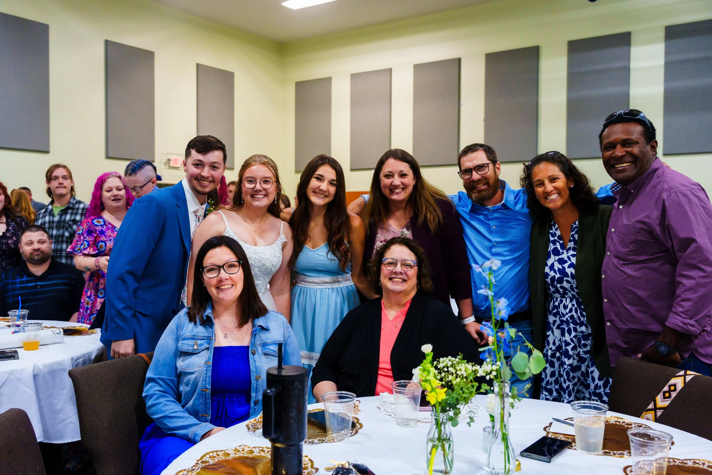 A group of diverse smiling people gathered at a celebration or event, some standing and some sitting at a table decorated with flowers, drinks, and tableware, in an indoor venue with gray soundproof panels on the walls.