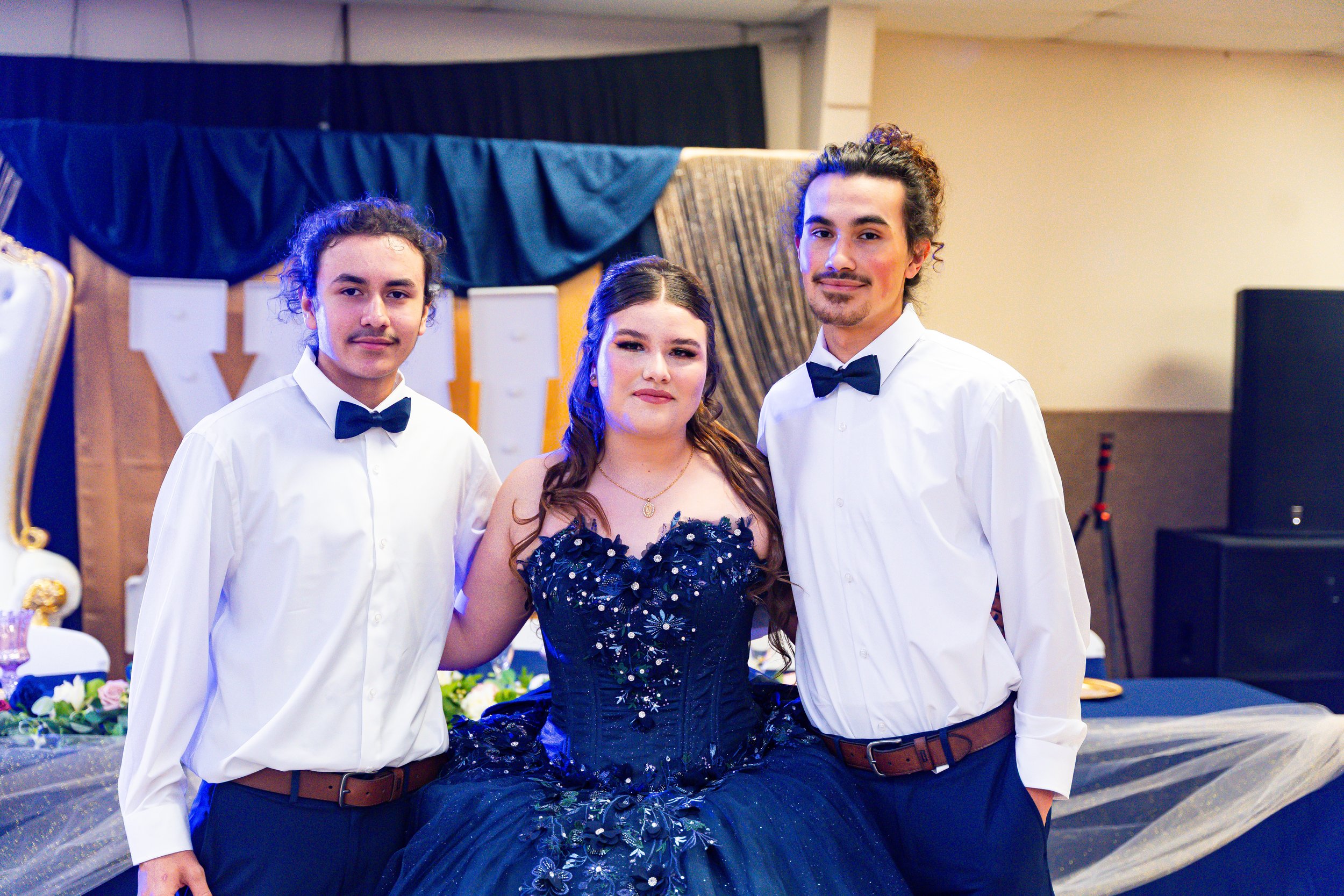 Three young adults dressed formally at a celebration or event, with a decorated backdrop and floral arrangements.