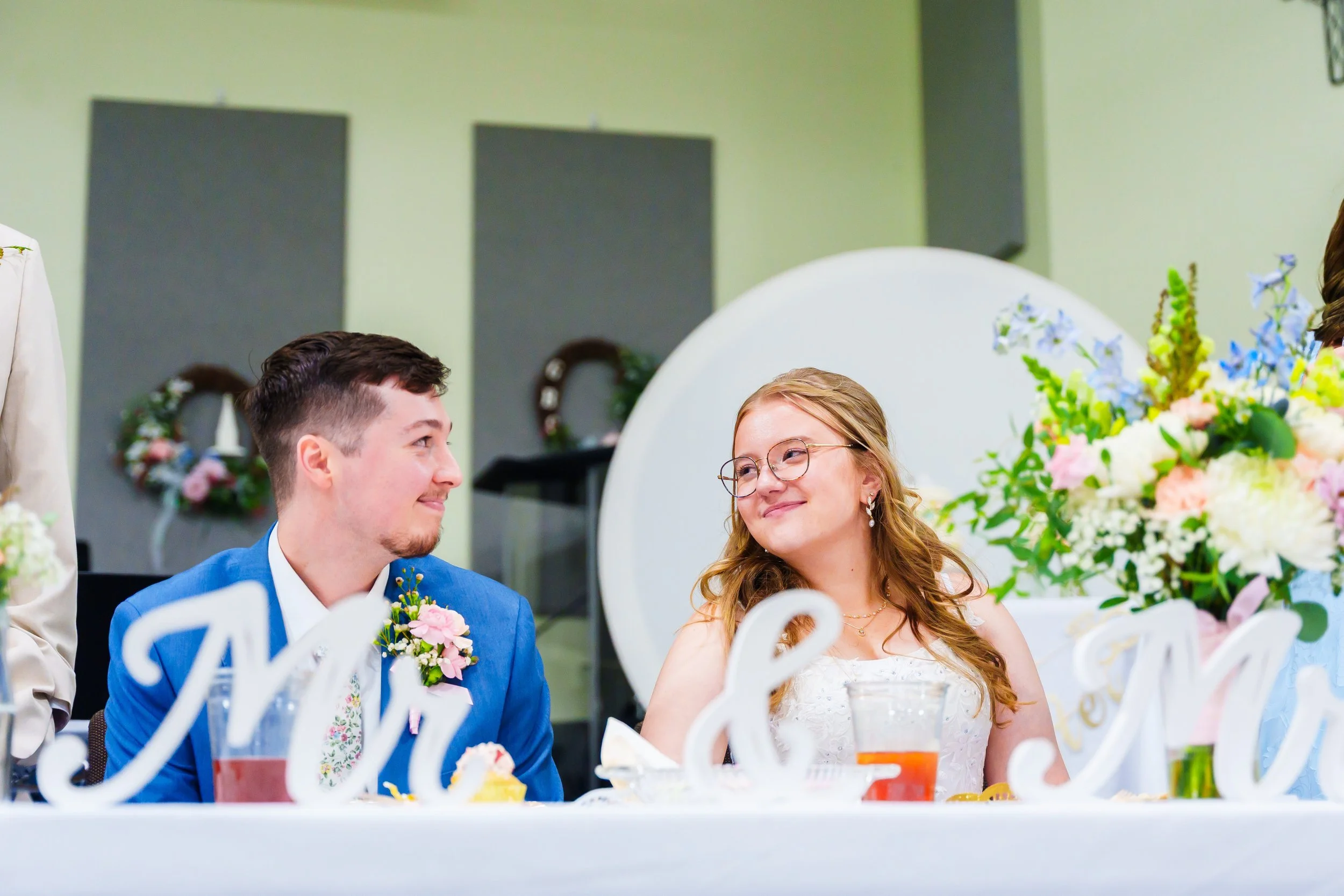 A bride and groom sitting at a table during their wedding reception, smiling and looking at each other. The table is decorated with flowers and wedding signs.