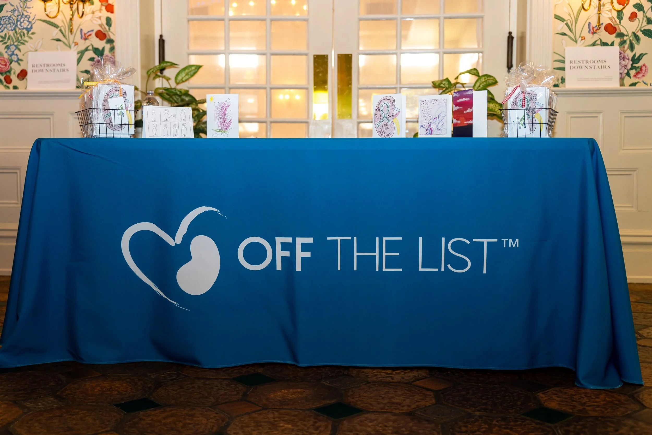 Table with blue tablecloth displaying promotional materials and gift baskets, with a stained glass window and floral wallpaper in the background.