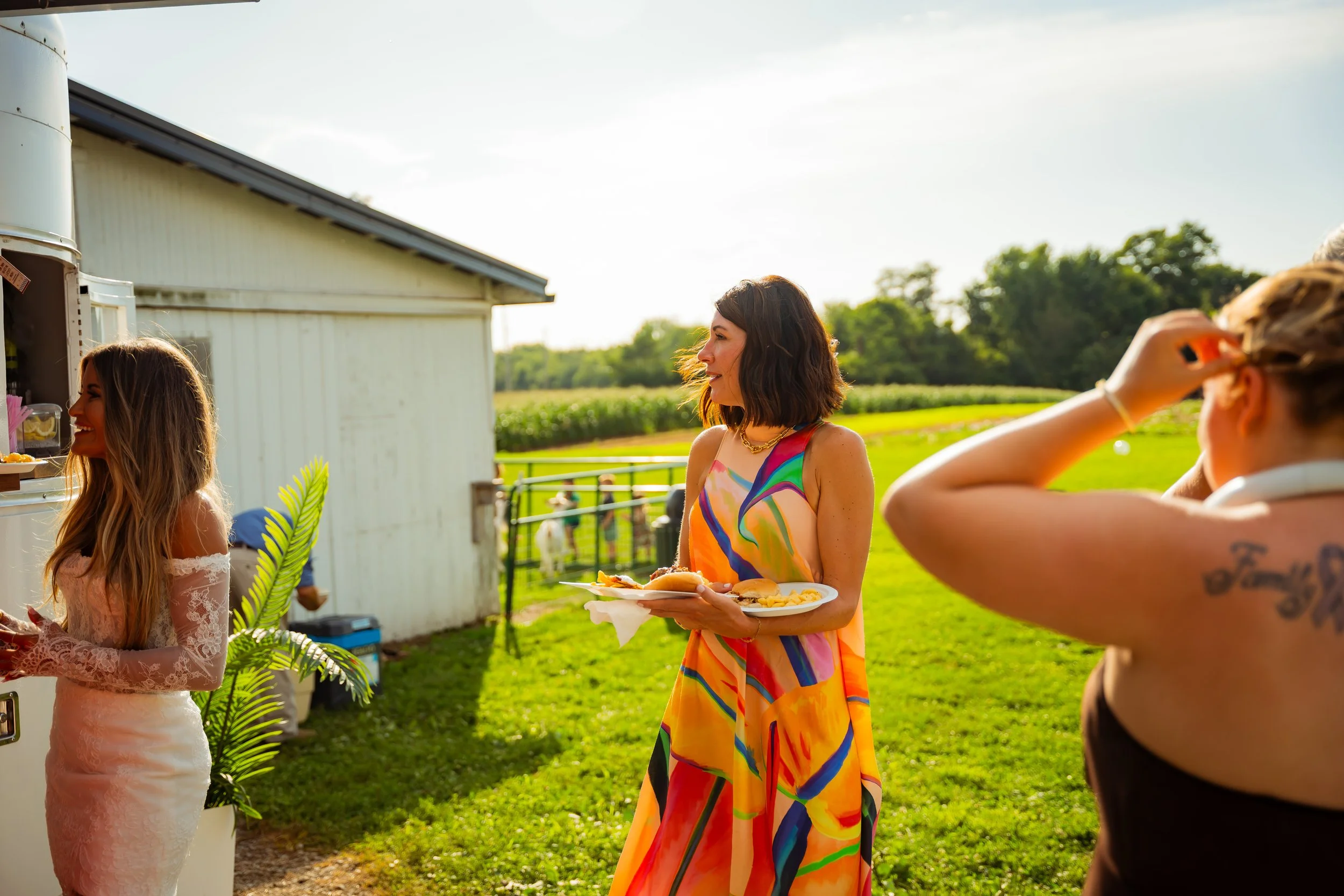People socializing outdoors on a sunny day, with a woman in a colorful dress holding a tray of food, and others taking photos or talking in the background near a farm or rural area.