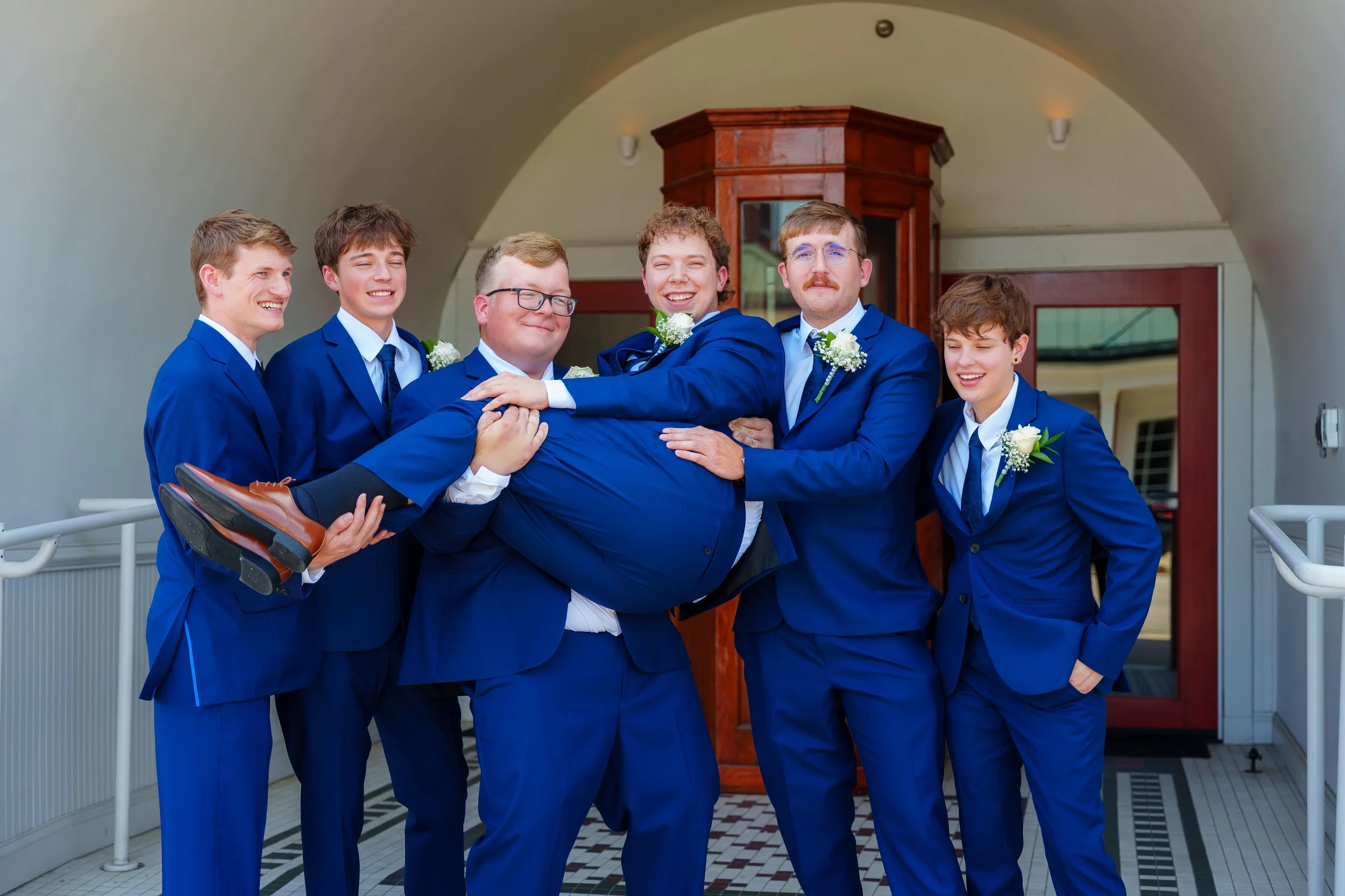 Group of six young adults in matching blue suits, with boutonnieres, posing playfully indoors, with one person being lifted horizontally by the others.