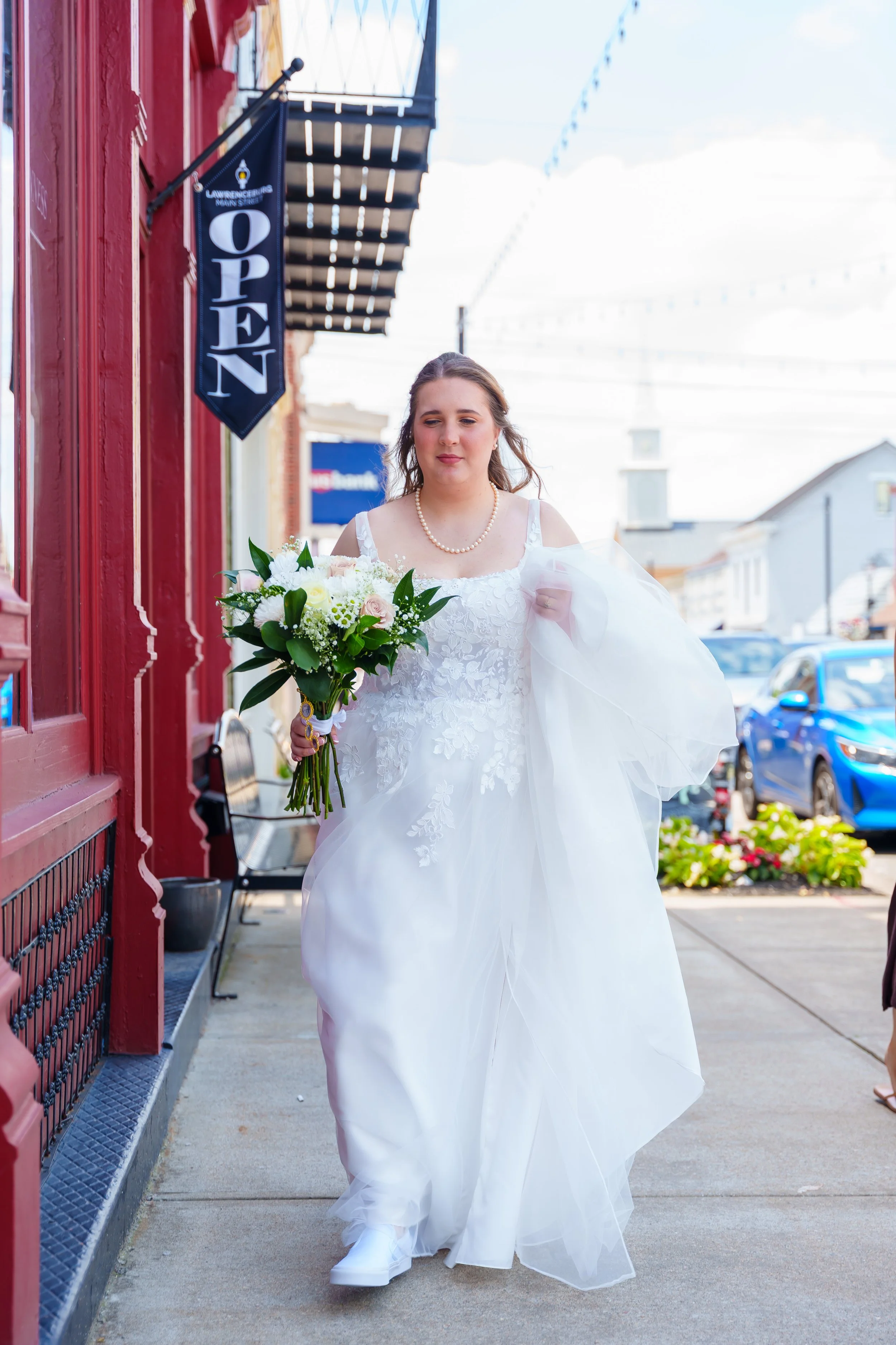 A bride in a white wedding dress holding a bouquet of flowers walking down a city sidewalk.