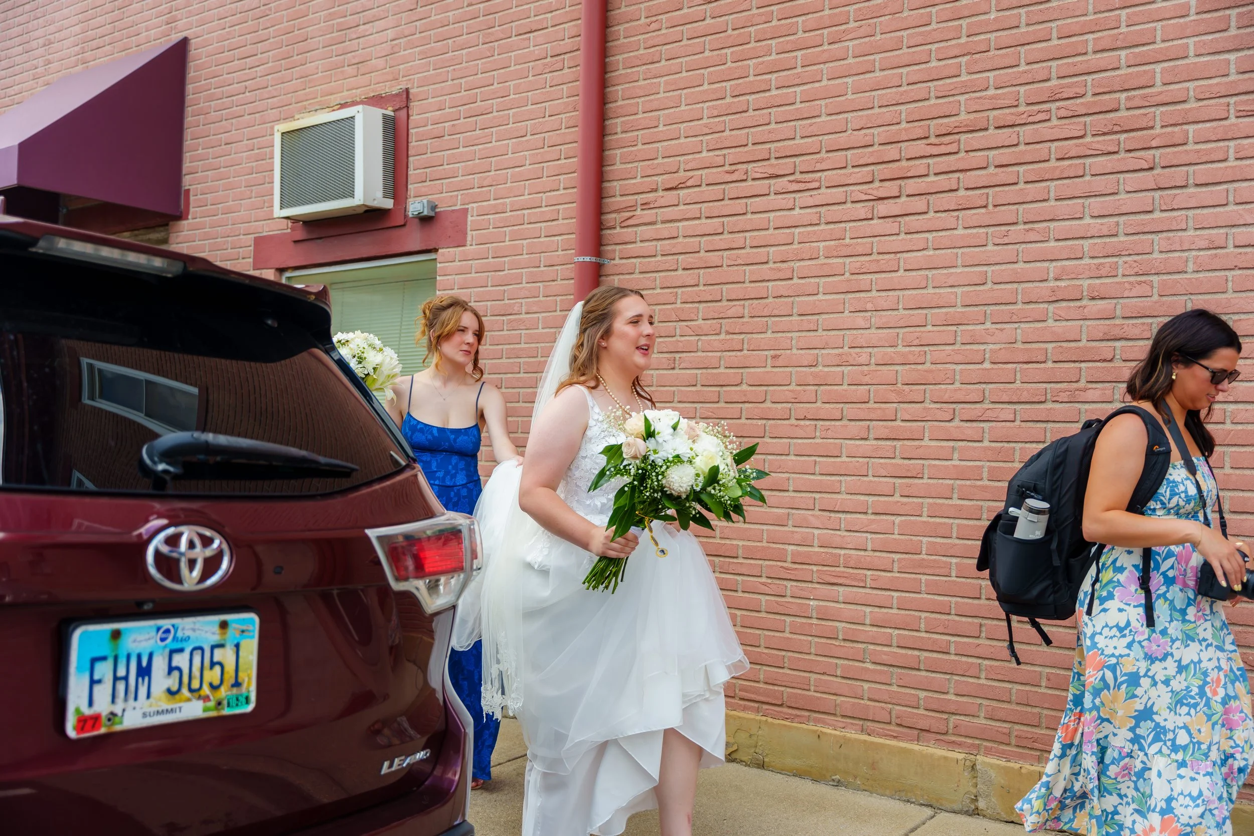 A bride wearing a white wedding dress and holding a bouquet of white flowers walking with three women, one in a blue dress, another in a floral dress and sunglasses, and a third woman behind the bride. They are outside next to a brick wall and a maro