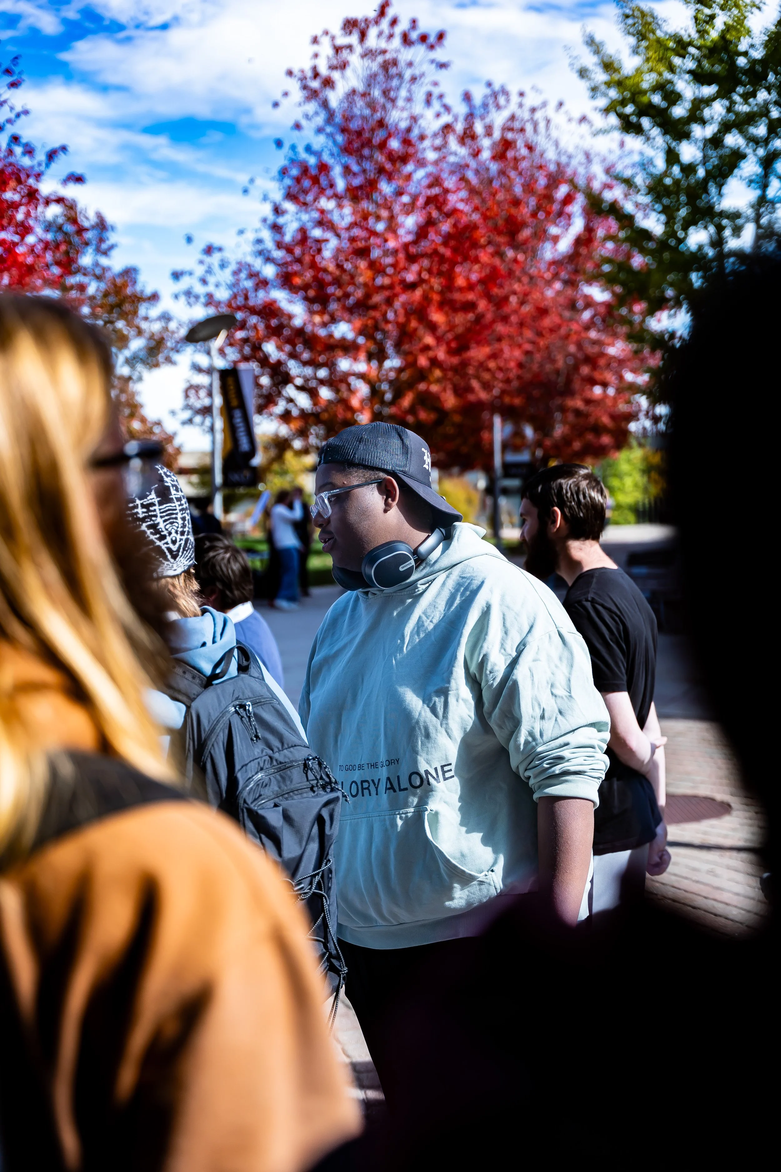 A group of people gathered outdoors during the daytime, with a bright red tree and a blue sky with clouds in the background, including a young man wearing glasses, a gray hoodie, a cap backward, and headphones around his neck.