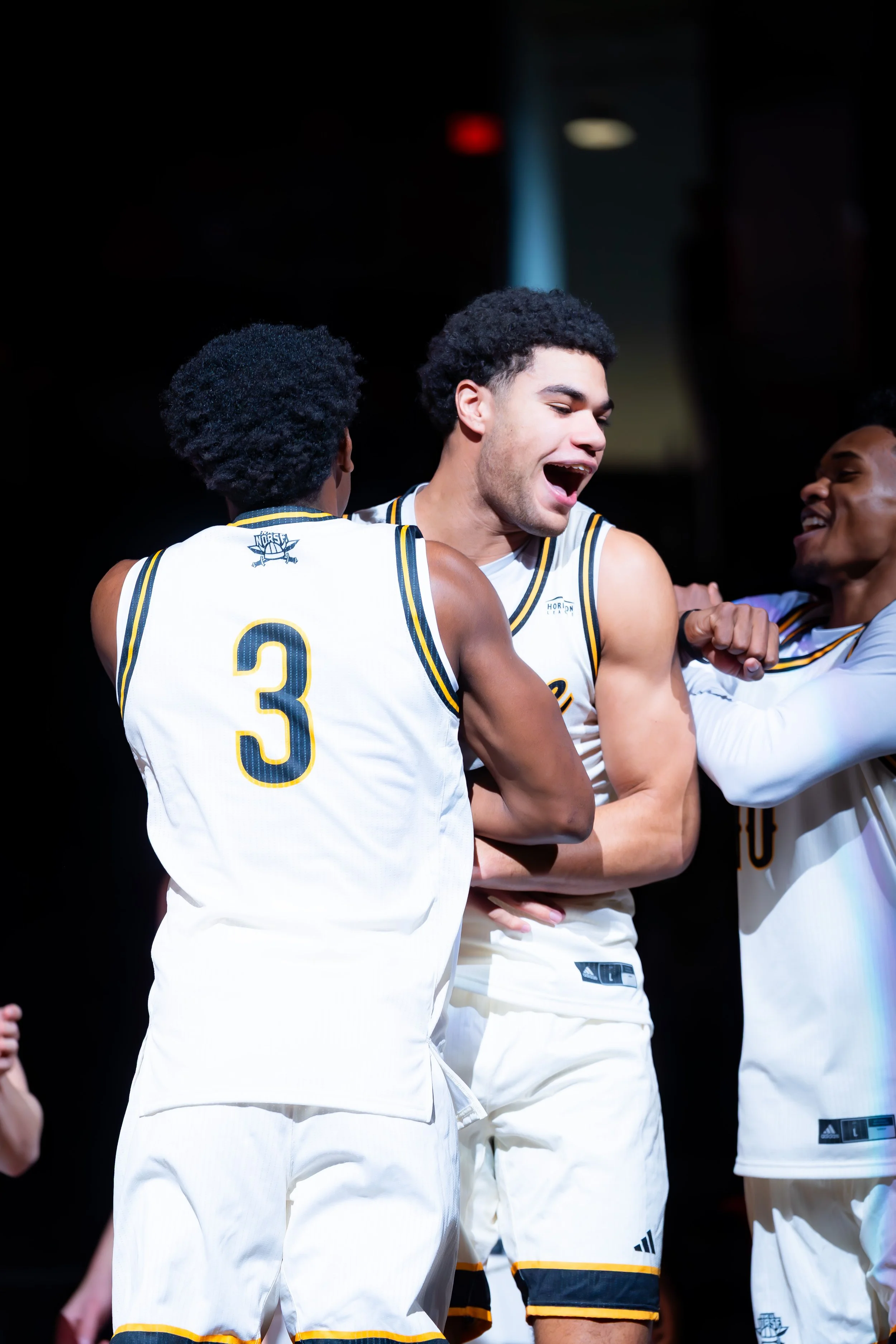 Three basketball players celebrating on the court, wearing white jerseys with navy and yellow trim, one with the number 3, smiling and engaging with each other.
