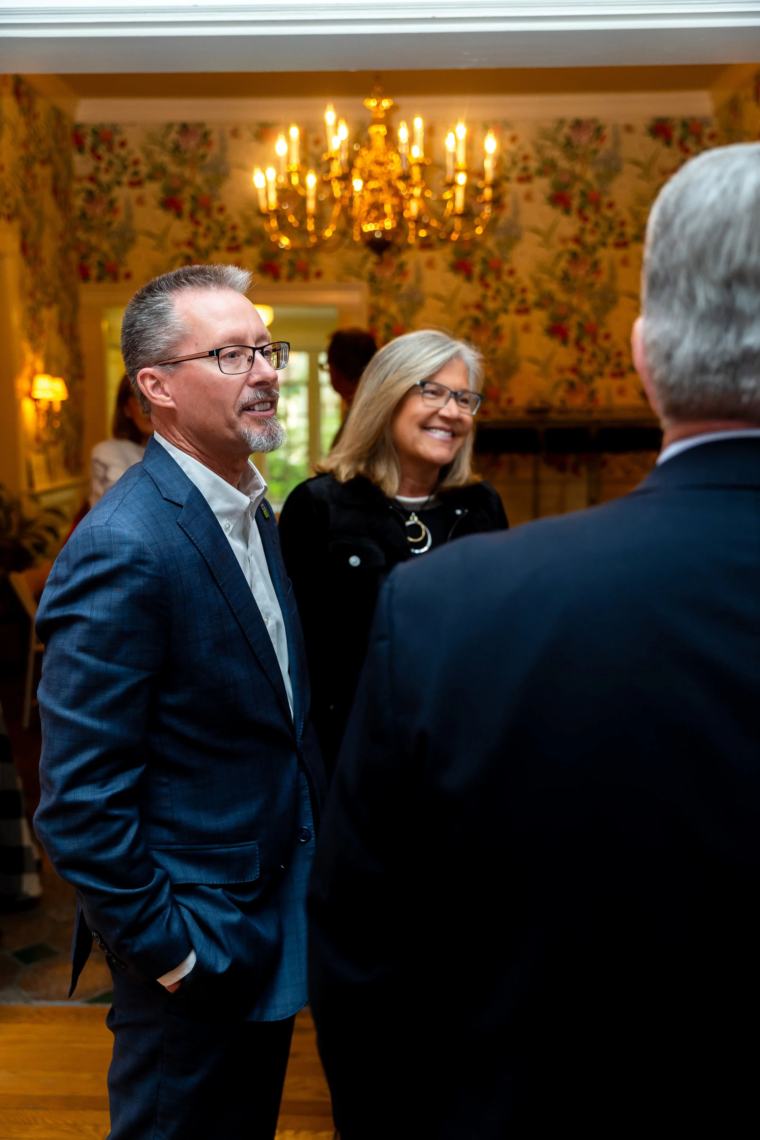Three people in conversation at an indoor event, with a chandelier and floral wallpaper in background.