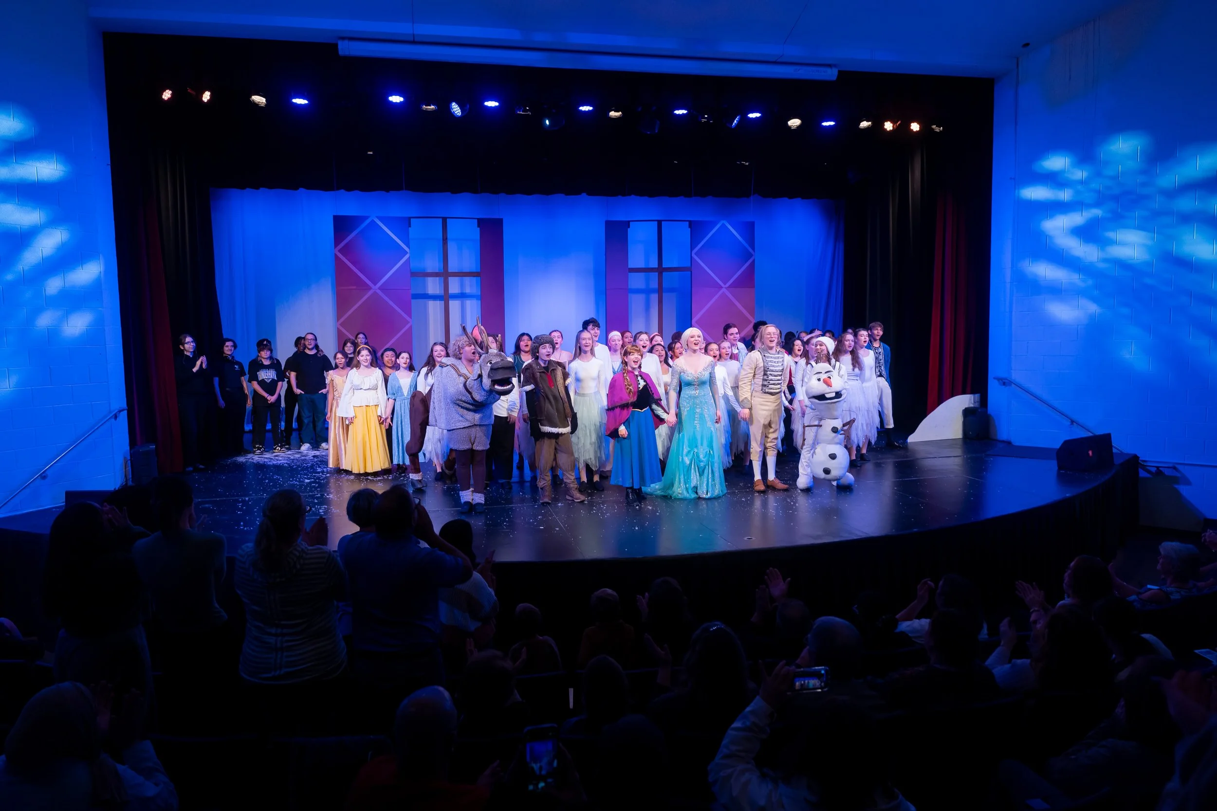 Group of children and performers dressed in costumes on stage after a theatrical performance, with an audience watching.