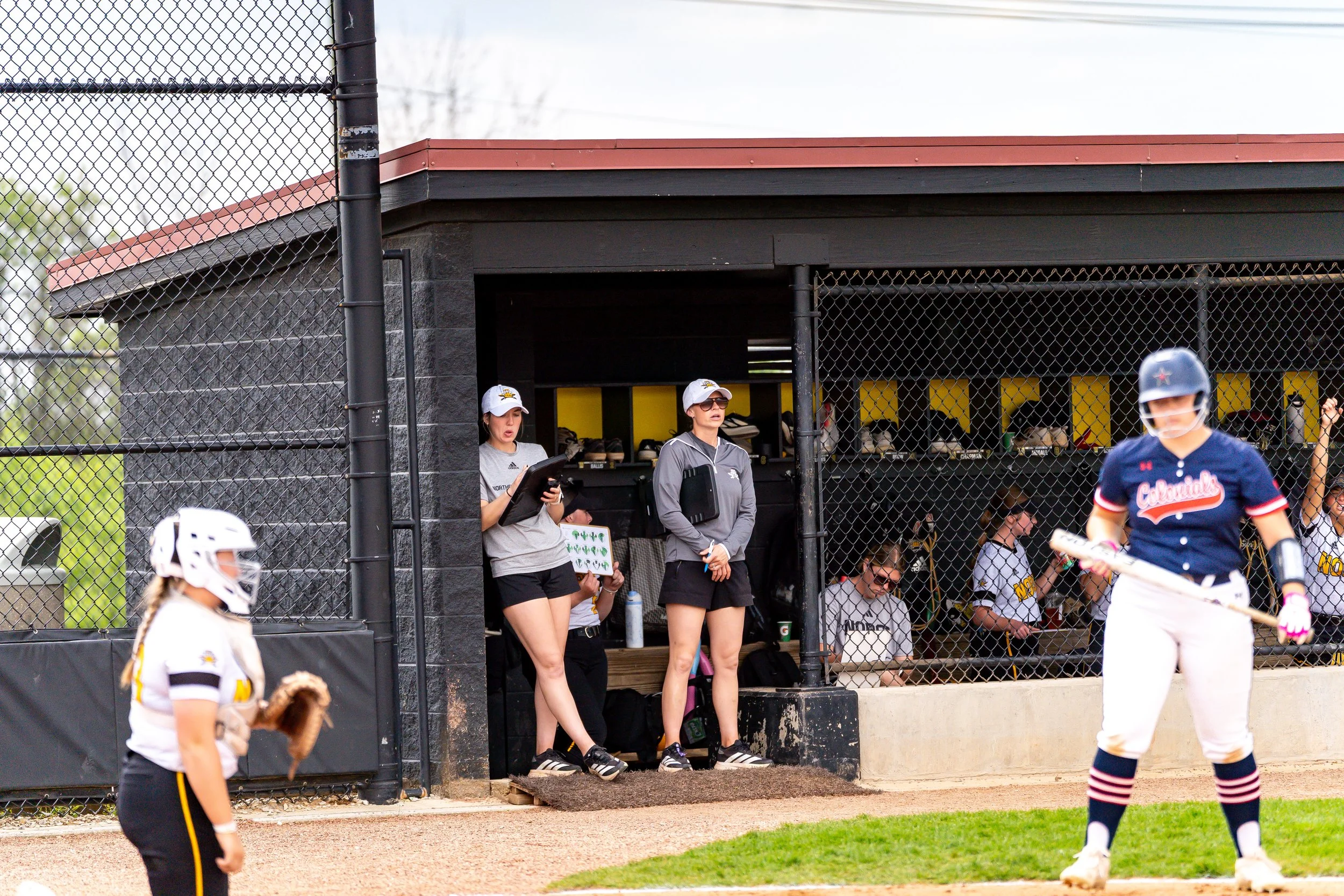 A softball game scene showing players on the field and coaches near the dugout, with a focus on a young player in a white and black uniform and helmet.