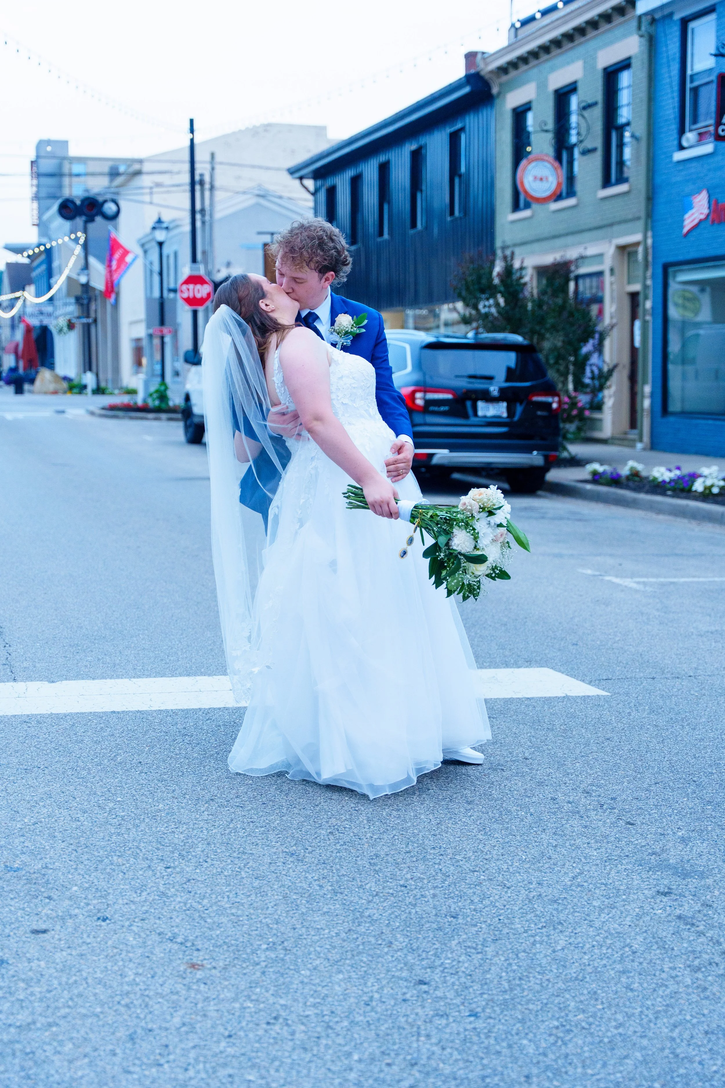 A newlywed couple sharing a kiss in the middle of a city street. The bride is wearing a white wedding gown and holding a bouquet of flowers, and the groom is dressed in a blue suit with a boutonniere. They are standing on the crosswalk with old build