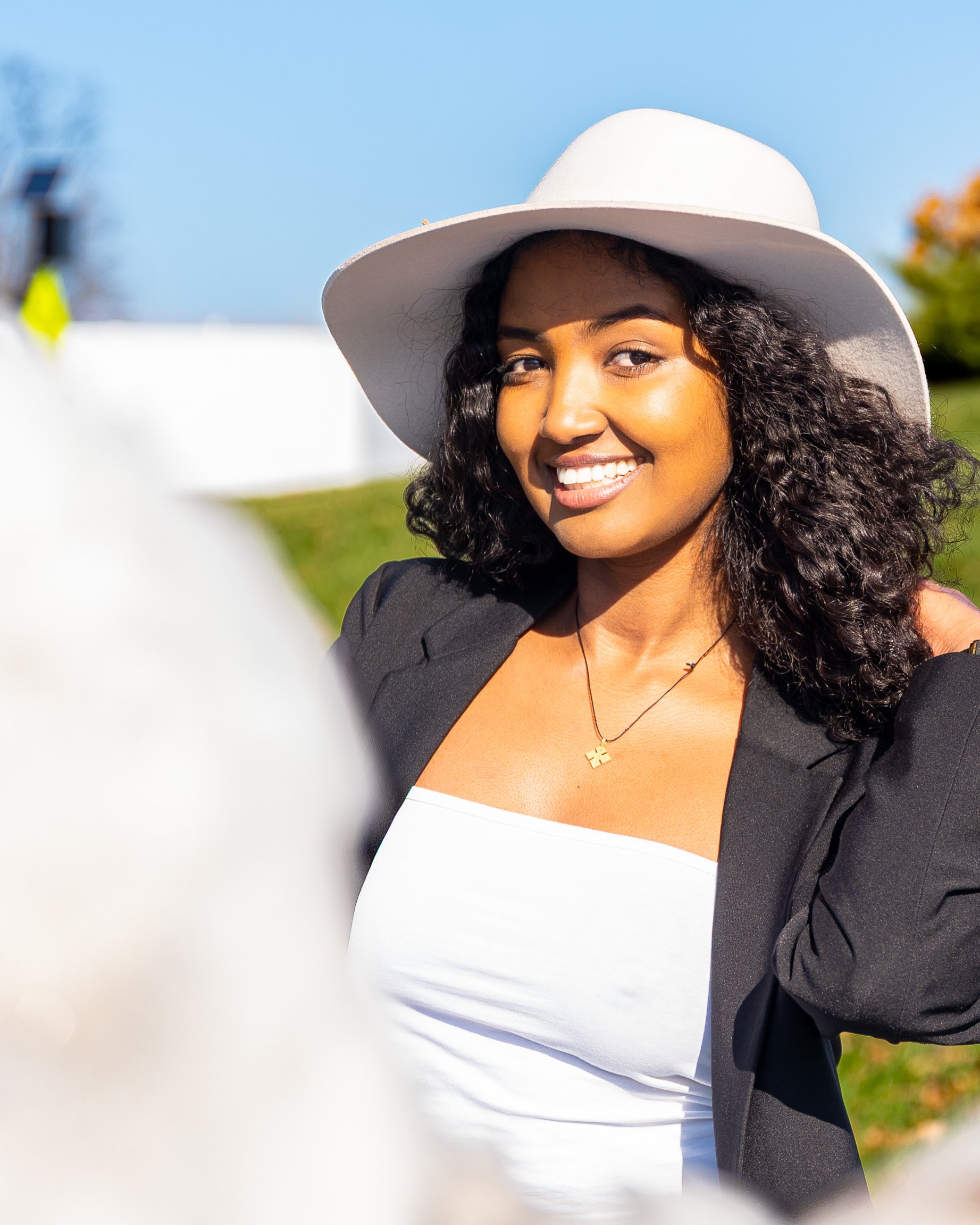 Smiling woman with curly hair in a white hat, black jacket, and white top outdoors on a sunny day.