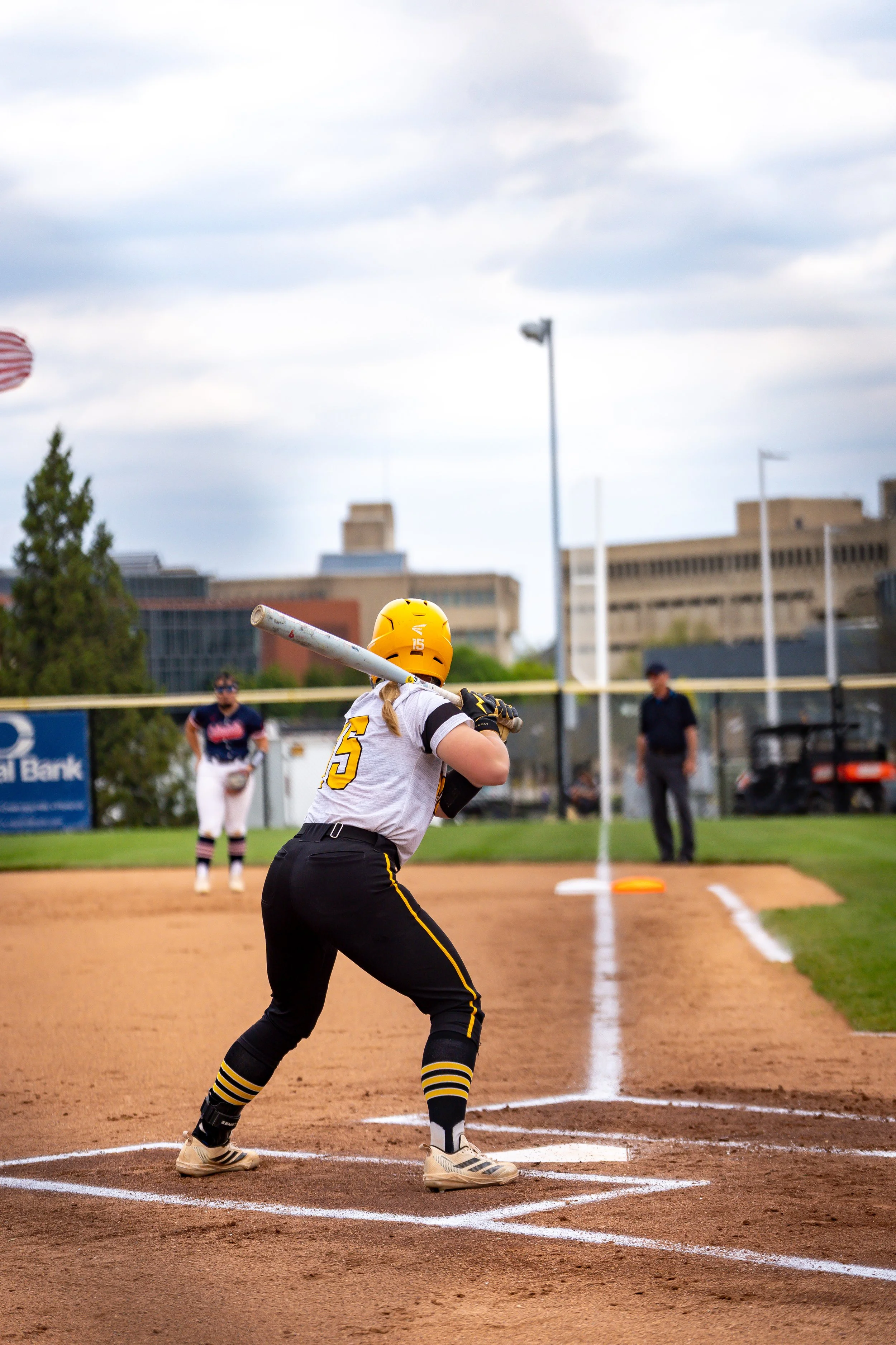 A female softball player wearing a yellow helmet and black uniform swinging a bat at a pitch on a softball field with a dirt infield and white chalk lines, with some players, spectators, and buildings in the background.