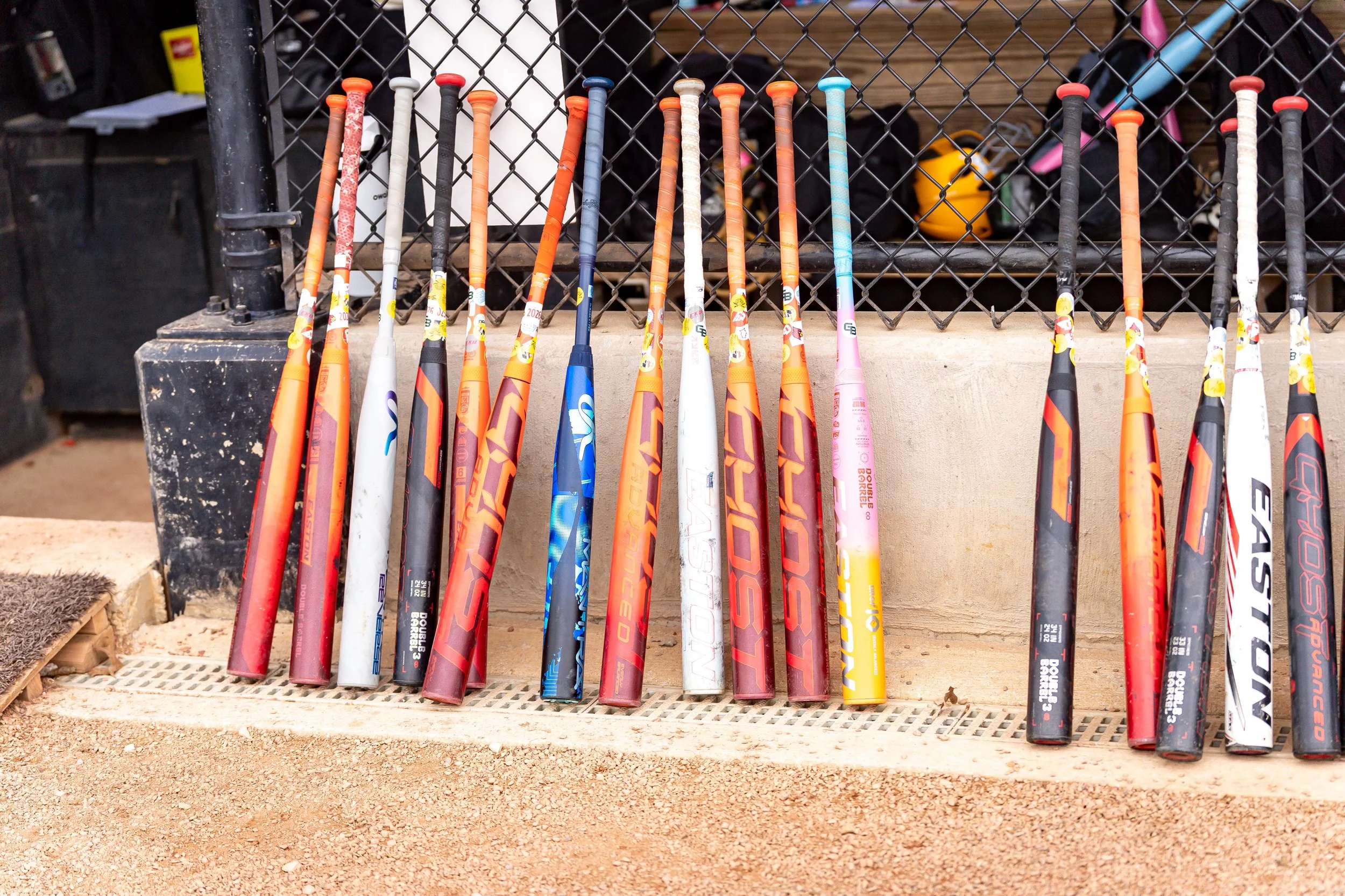 A row of colorful baseball bats leaning against a black metal fence, with various tools and equipment in the background.
