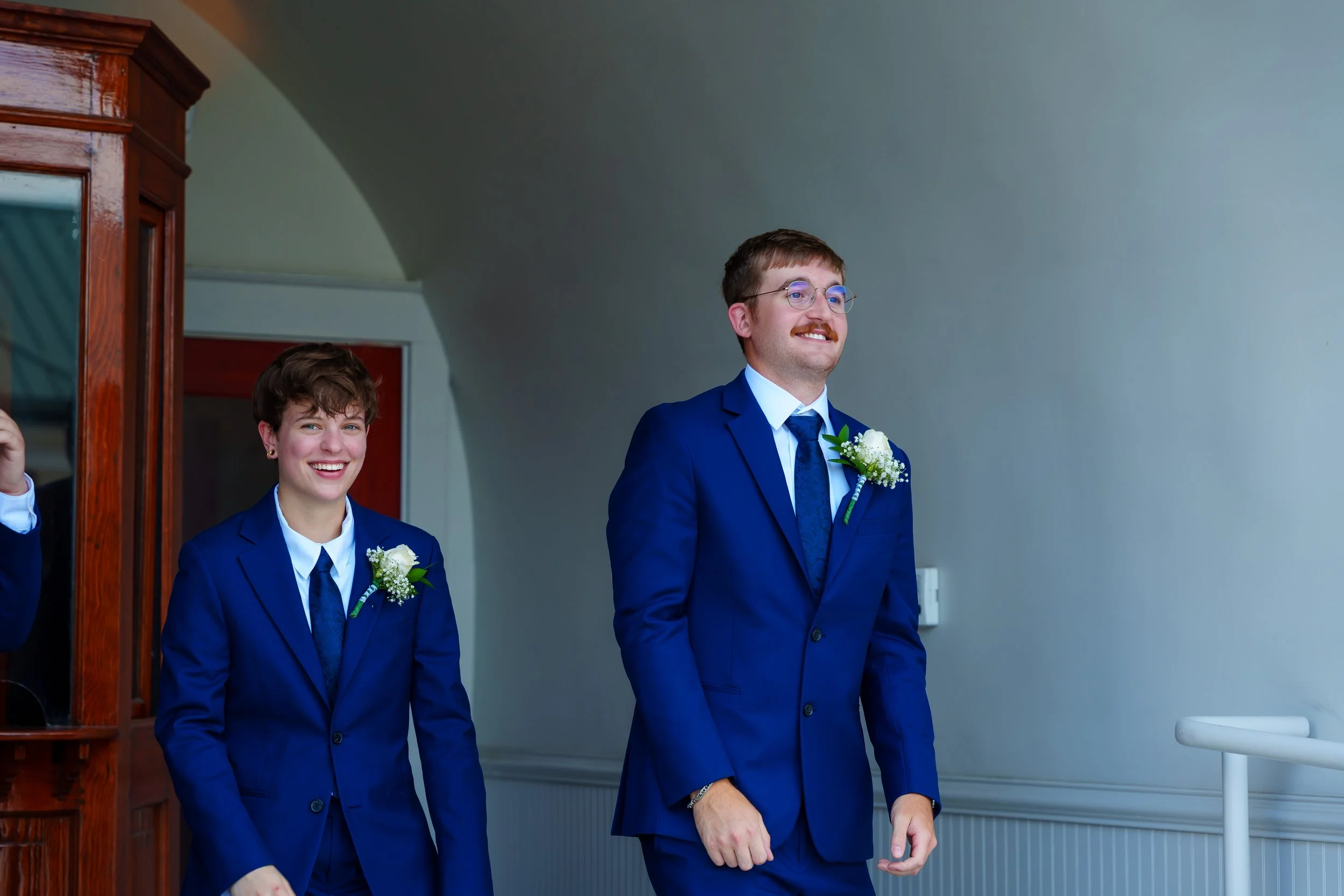 A man and a woman dressed in blue suits with white shirts and floral boutonnieres, standing indoors with a plain background. The man has glasses and a mustache, while the woman has short brown hair. They appear to be at a formal event or celebration,