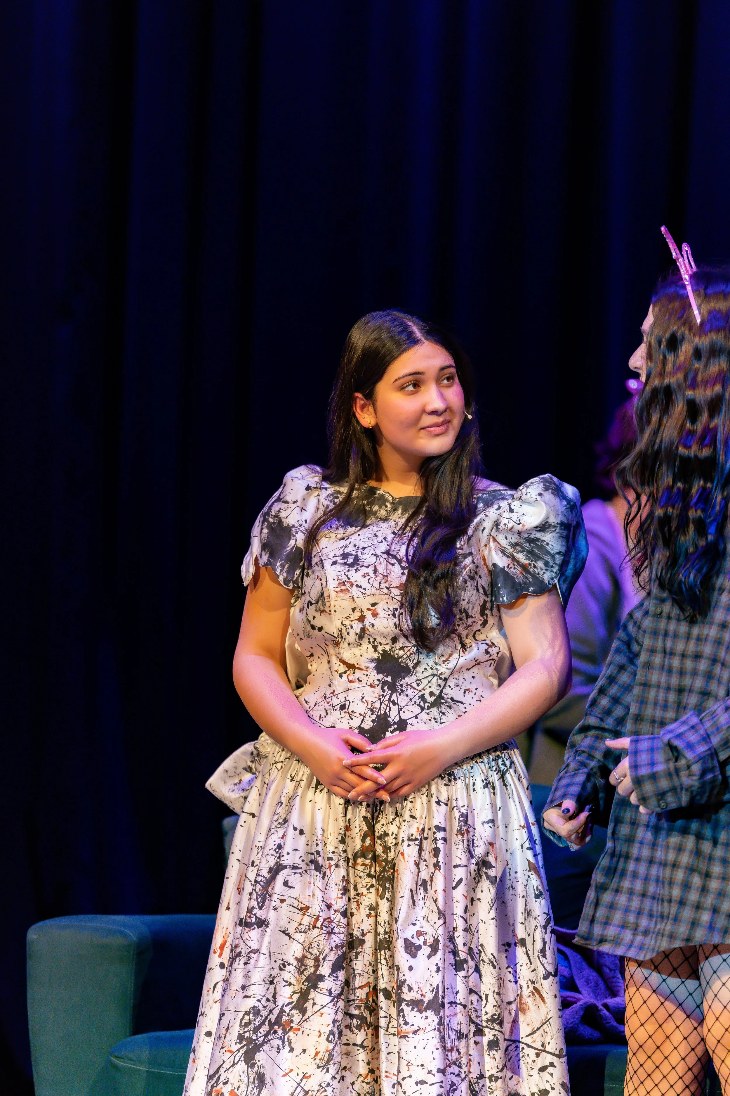 Two women on stage in conversation, one in a patterned dress with a floral print and the other in a checkered jacket and fishnet stockings, against a dark curtain backdrop.