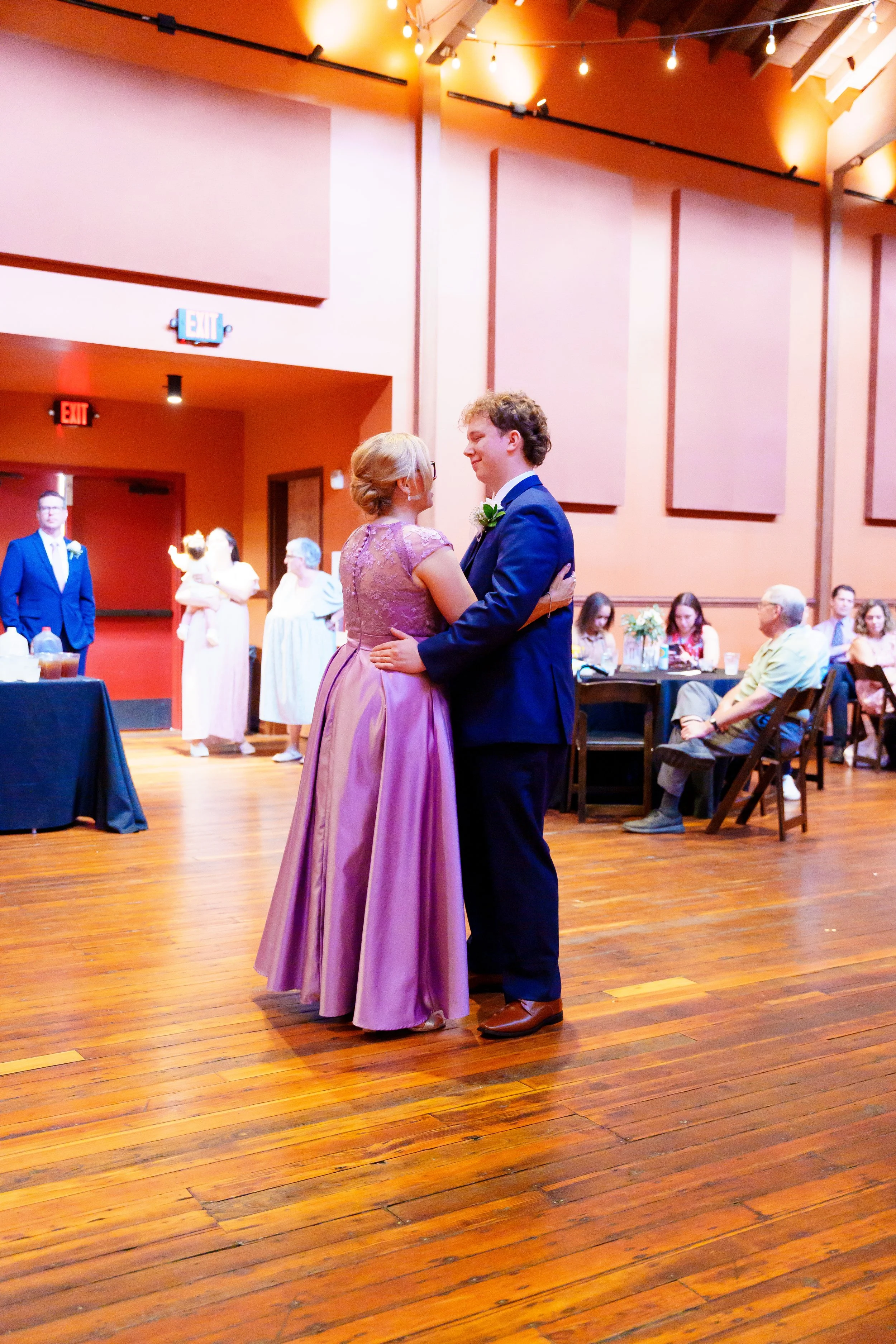 A bride and groom dance at their wedding reception, with guests seated at tables in the background and a woman holding a child to the left.