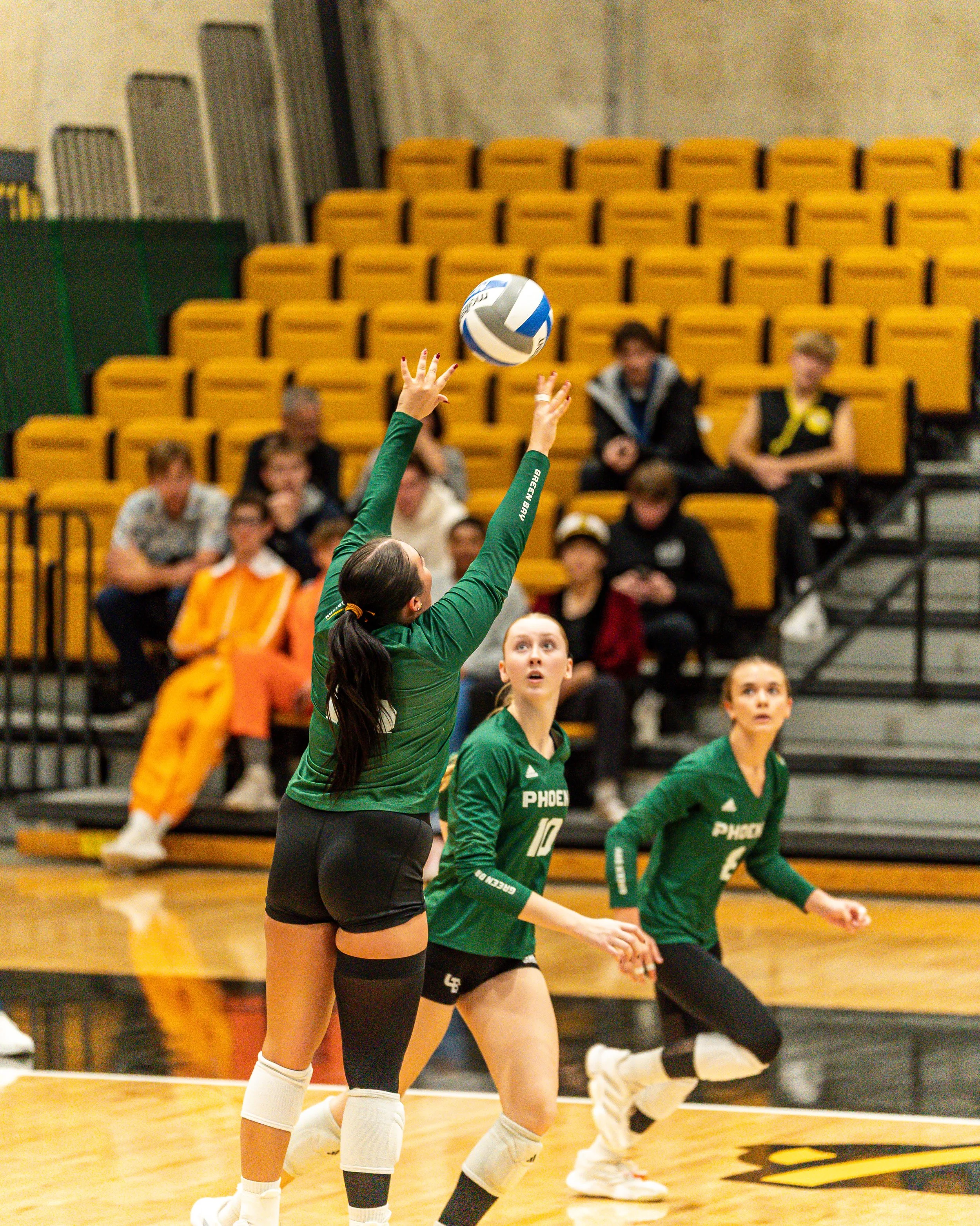 A women's volleyball game in progress with a player in green uniform jumping to hit the ball while another teammate prepares to assist. Spectators are seated in yellow bleachers in the background.