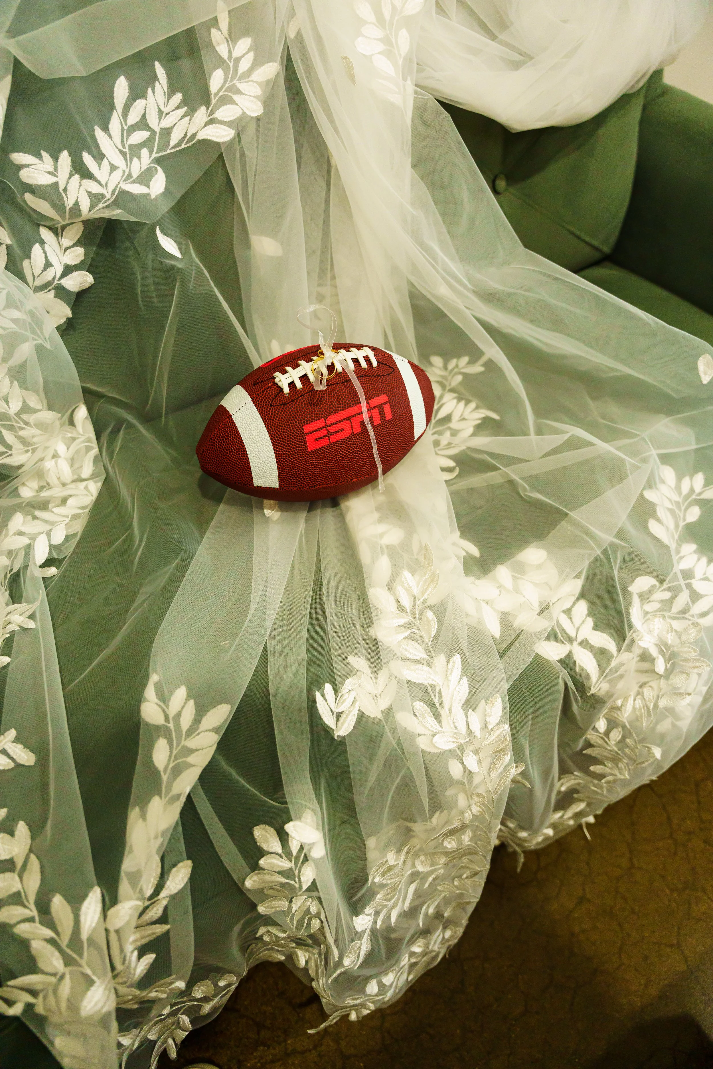 A decorative green tablecloth with white embroidered leaf patterns. On the tablecloth, there is a red and white American football with the ESPN logo on it, placed amidst some sheer fabric with floral embroidery.